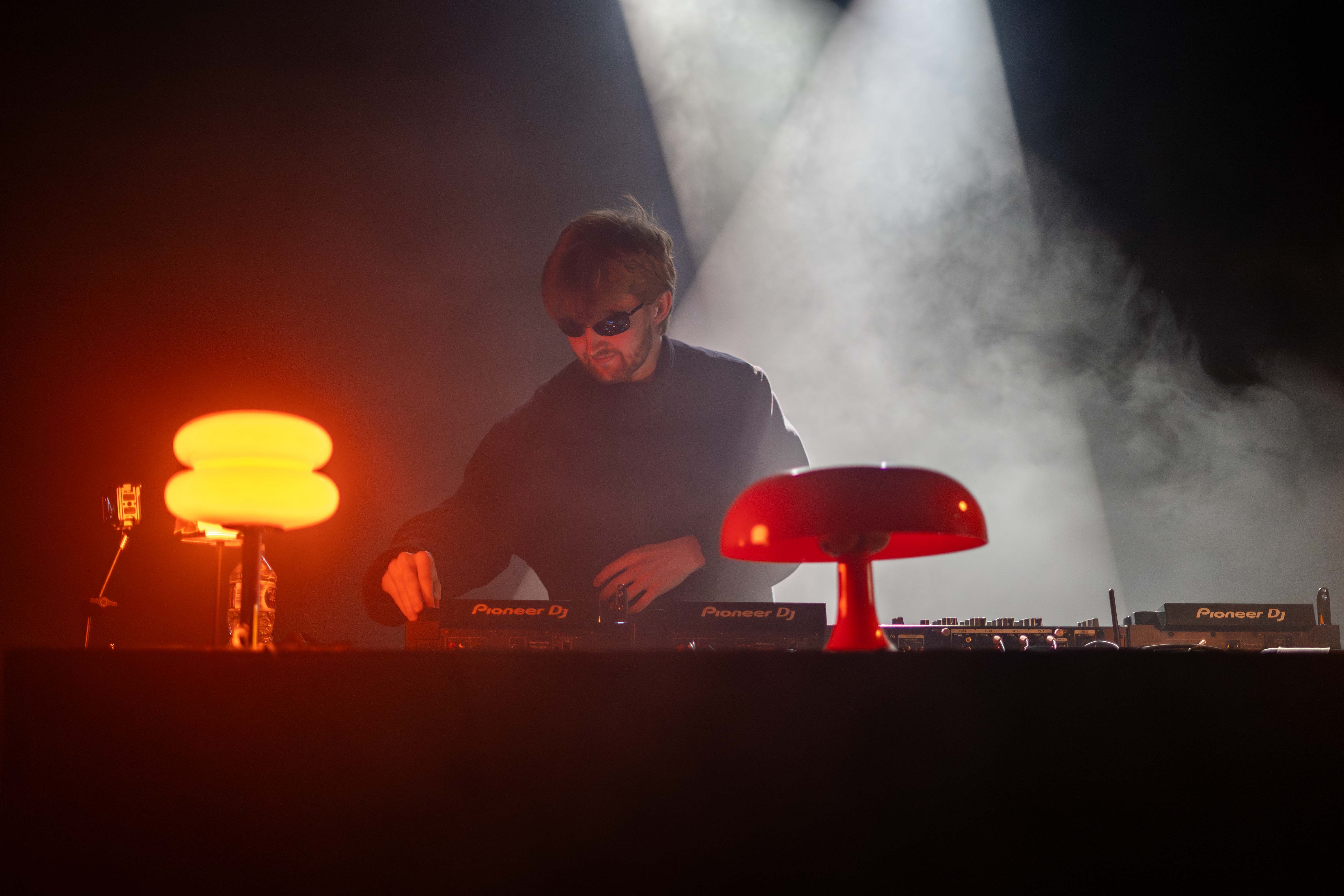 Oskar Med K is standing in front and adjusting a DJ deck and lamps wearing sunglasses with spotlights behind him