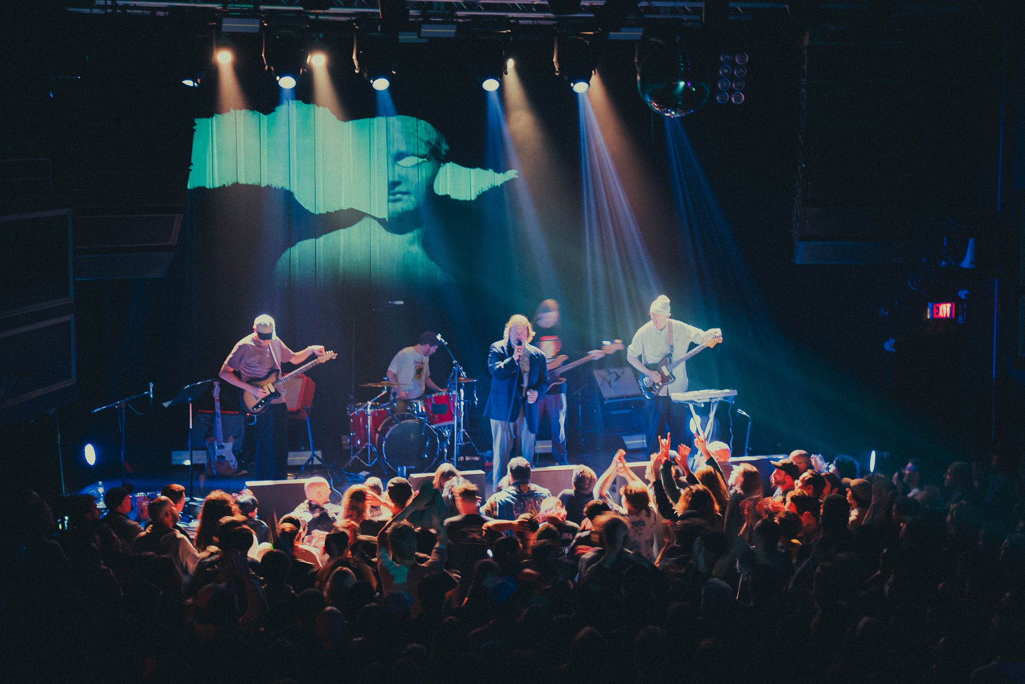Wide angle photo looking over the crowd towards the band playing on stage. An image of an ancient greek face sculpture is projected onto the wall behnd the band. The crowd is washed in warm light as subtle motion blur shows their moshing and dancing. A few crowd members have their fists in the air making "rock and roll" gestures.