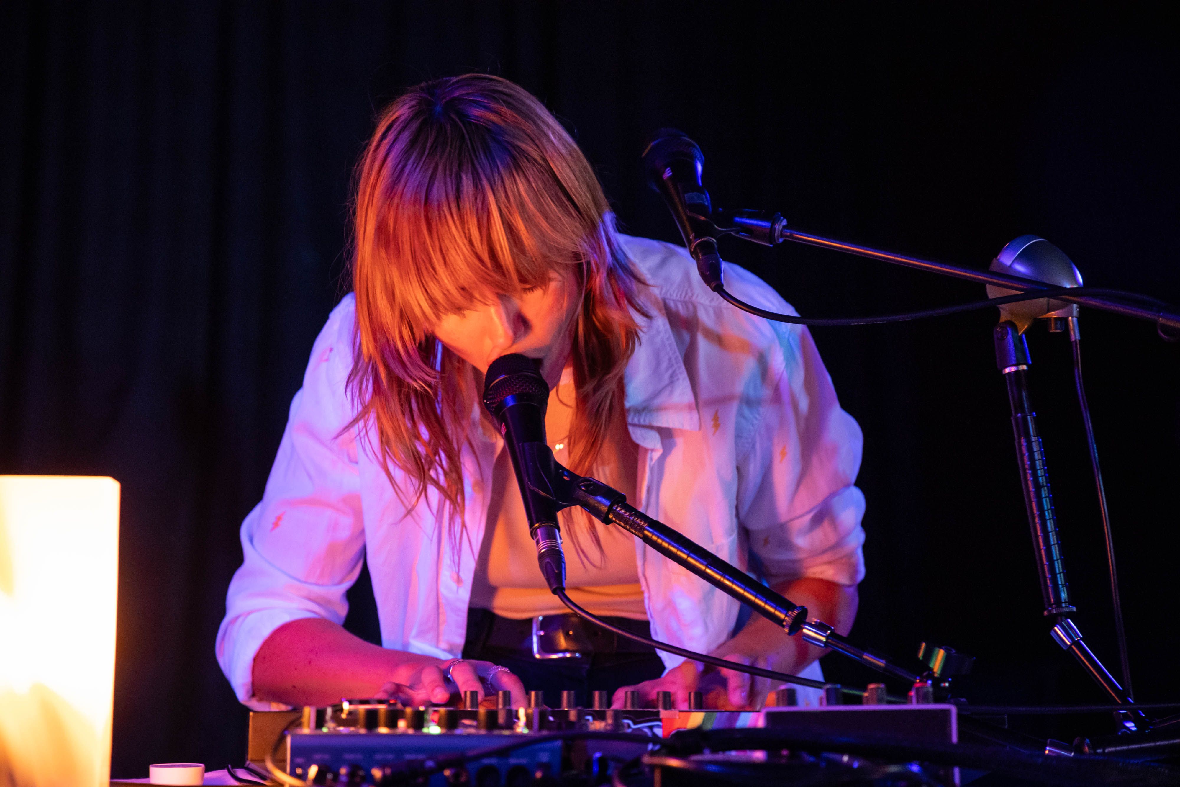 Gordi leans over her keyboard to sing into an FX mic, lit by warm lighting