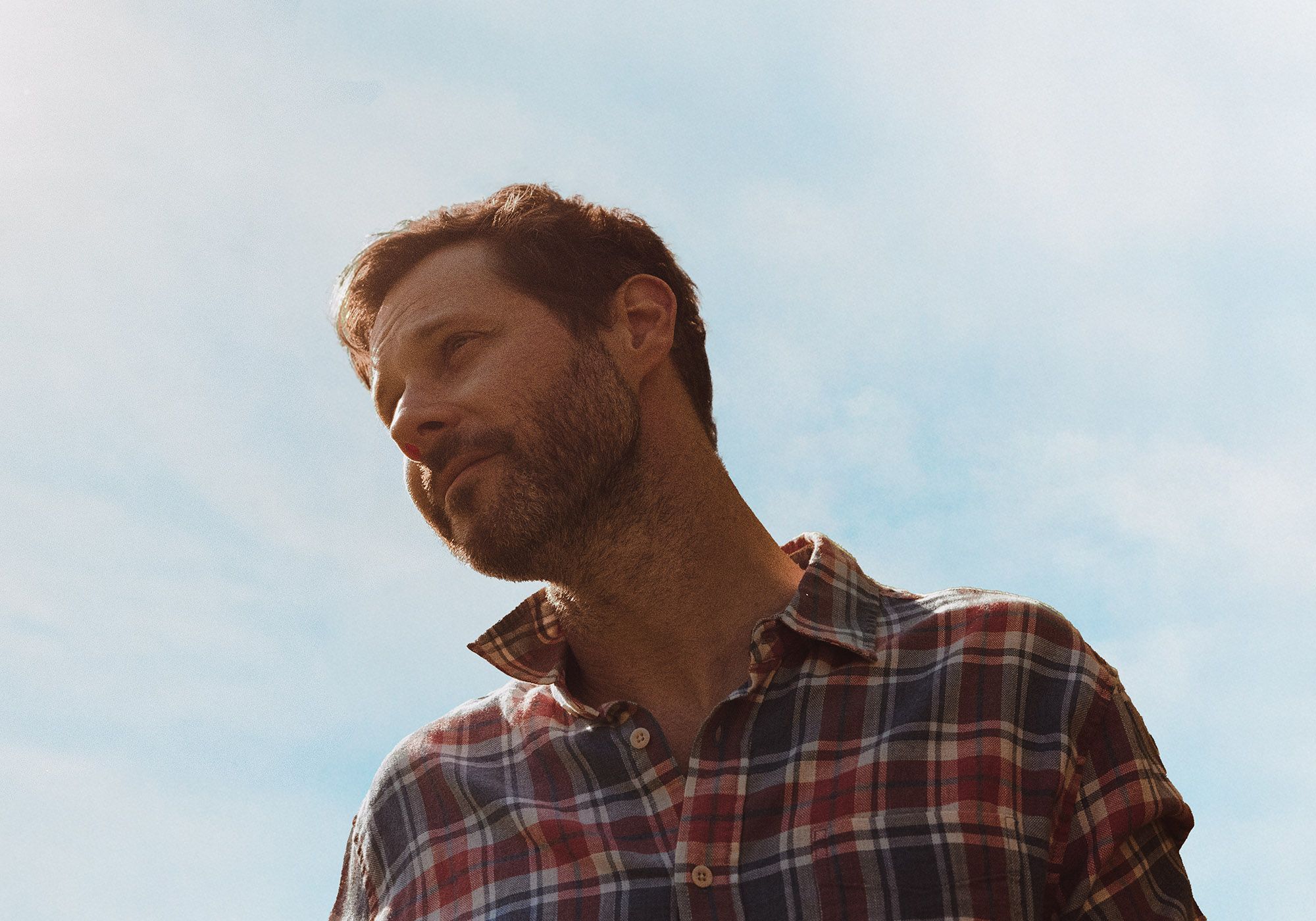 Dan Mangan wearing a plaid shirt against a background of blue sky.