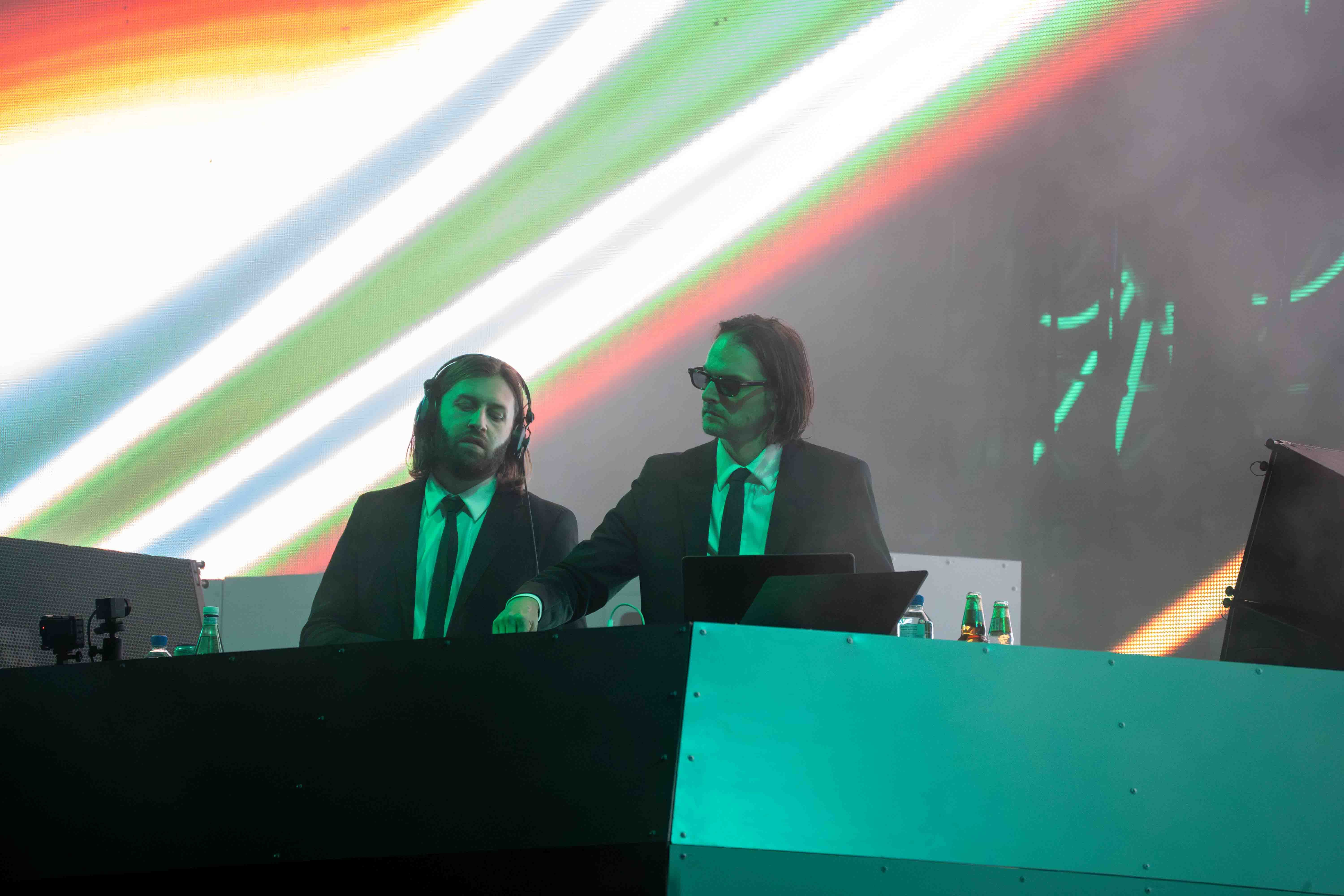 Zeds Dead standing behind a DJ deck in a black suit and tie with sunglasses adjusting a knob on the Deck with colourful visuals behind them.