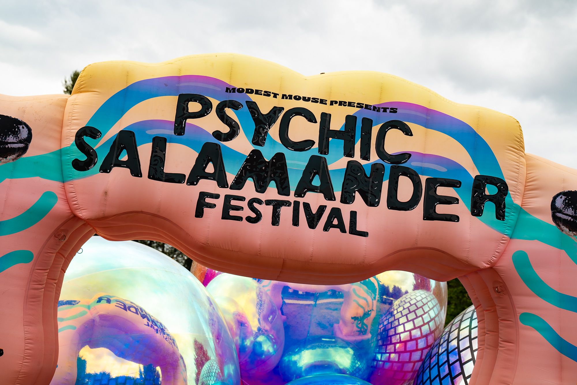 A brightly coloured inflatable archway with "Modest Mouse presents Psychic Salamander Festival" printed in large block letters and adorned with iridescent balloons welomes guests to the festival grounds.
