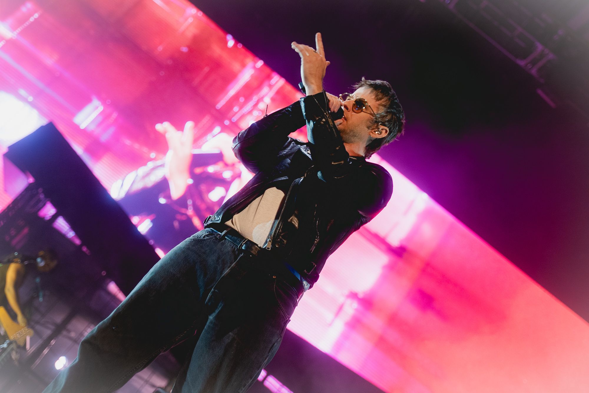 Mark Foster singing into a microphone and posing in a dance movement in front of a large LED screen and scaffolding.