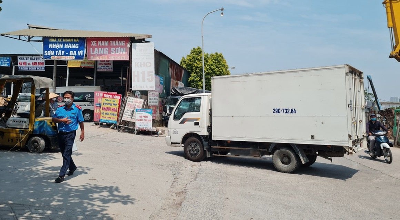 Elevated view of a Vietnamese logistics warehouse district, Northern Vietnam industrial area, corrugated metal warehouses, delivery trucks lined up at loading docks, a wide access road with trucks and motorcycles, flat terrain with distant cityscape, bright sunny day, clear sky, natural colors, balanced white balance, clean natural light, documentary photography style