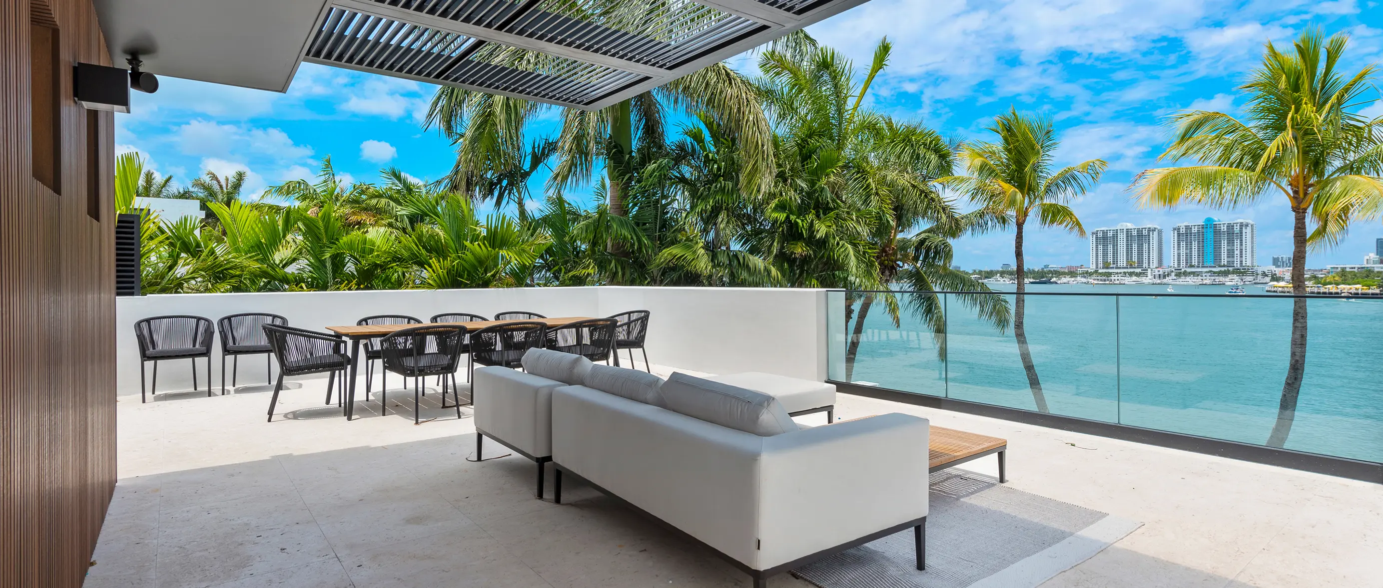 Covered outdoor patio with modern furnishings overlooking turquoise waterfront and palm trees