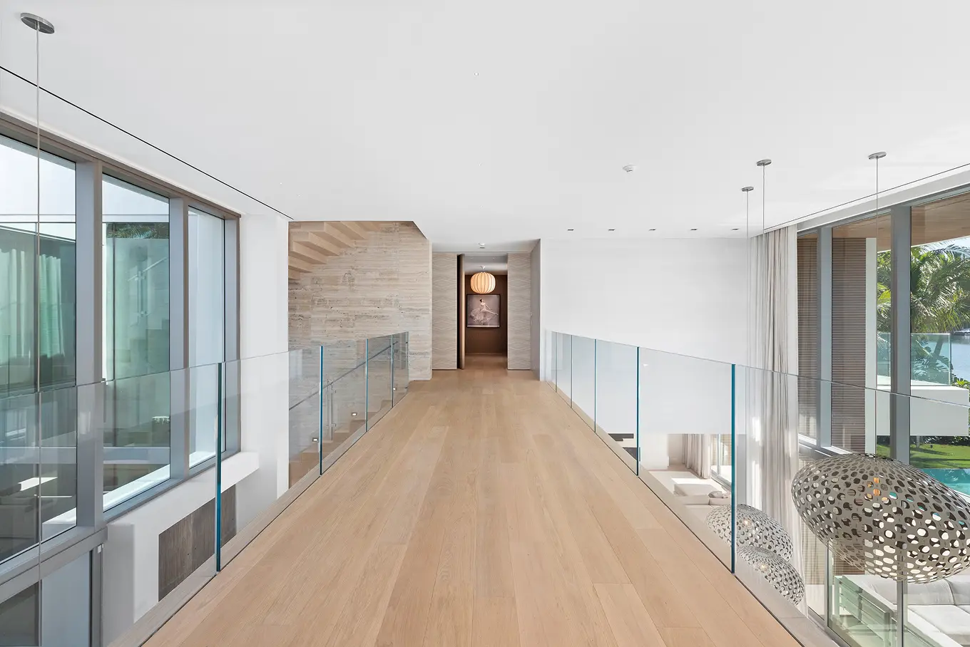 Second-floor bridge hallway with glass railings overlooking double-height living room and featuring warm wood accents