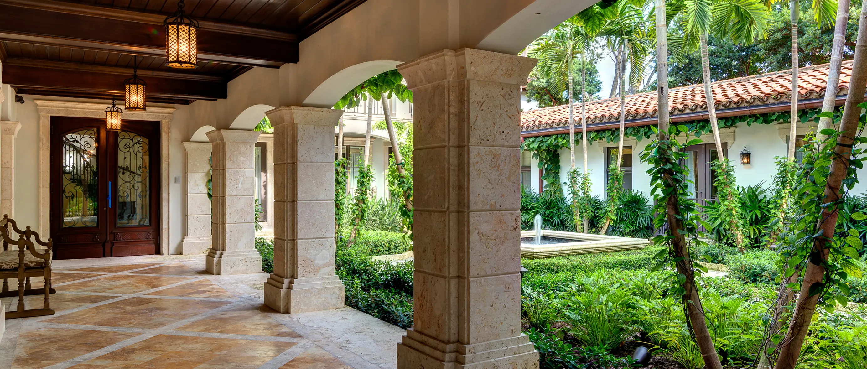 Covered colonnade with stone pillars and dark wood ceiling overlooking courtyard fountain and tropical gardens
