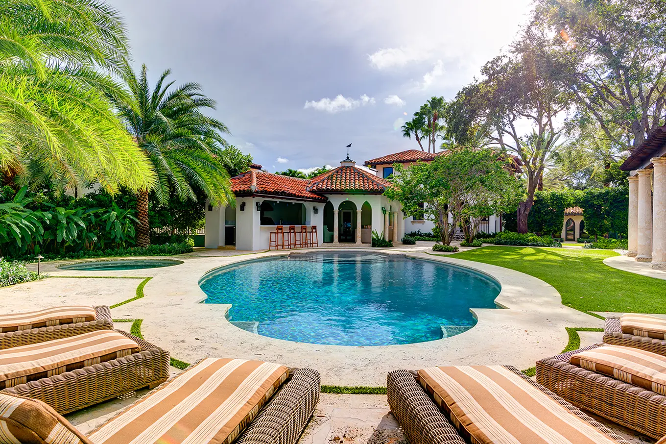 Resort-style pool with Spanish Colonial poolhouse cabana, outdoor bar, striped lounge seating, and lush tropical landscaping