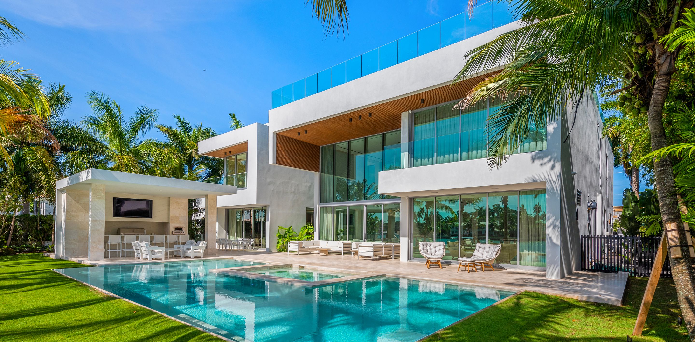 Modern waterfront estate with two-story glass facade, infinity pool, outdoor pavilion, and lush tropical landscaping on Miami bay