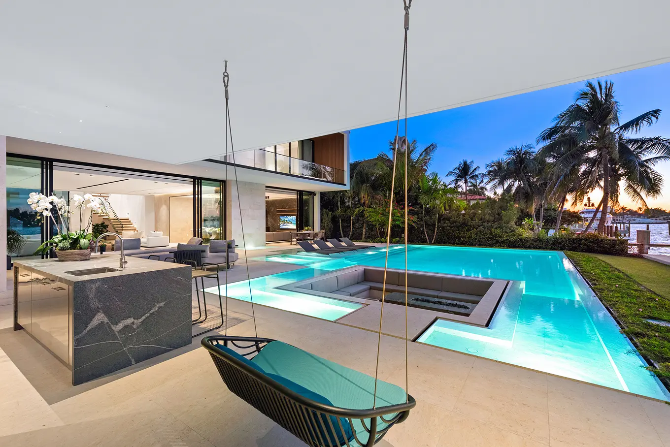 Stunning pool design with integrated fire pit lounge platform surrounded by illuminated water at night