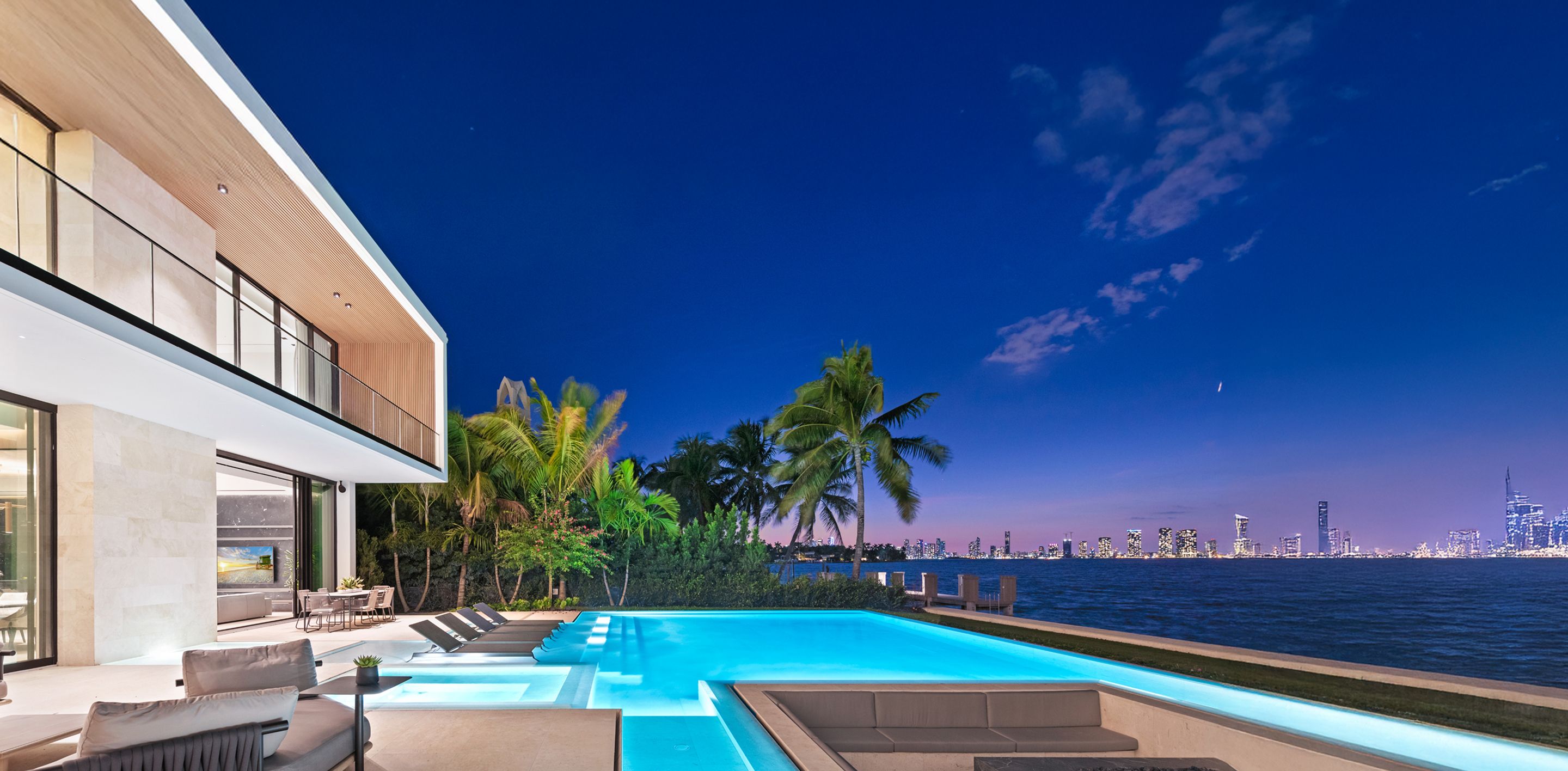 Modern waterfront estate at twilight with infinity pool, palm trees, and panoramic Miami skyline views across the bay