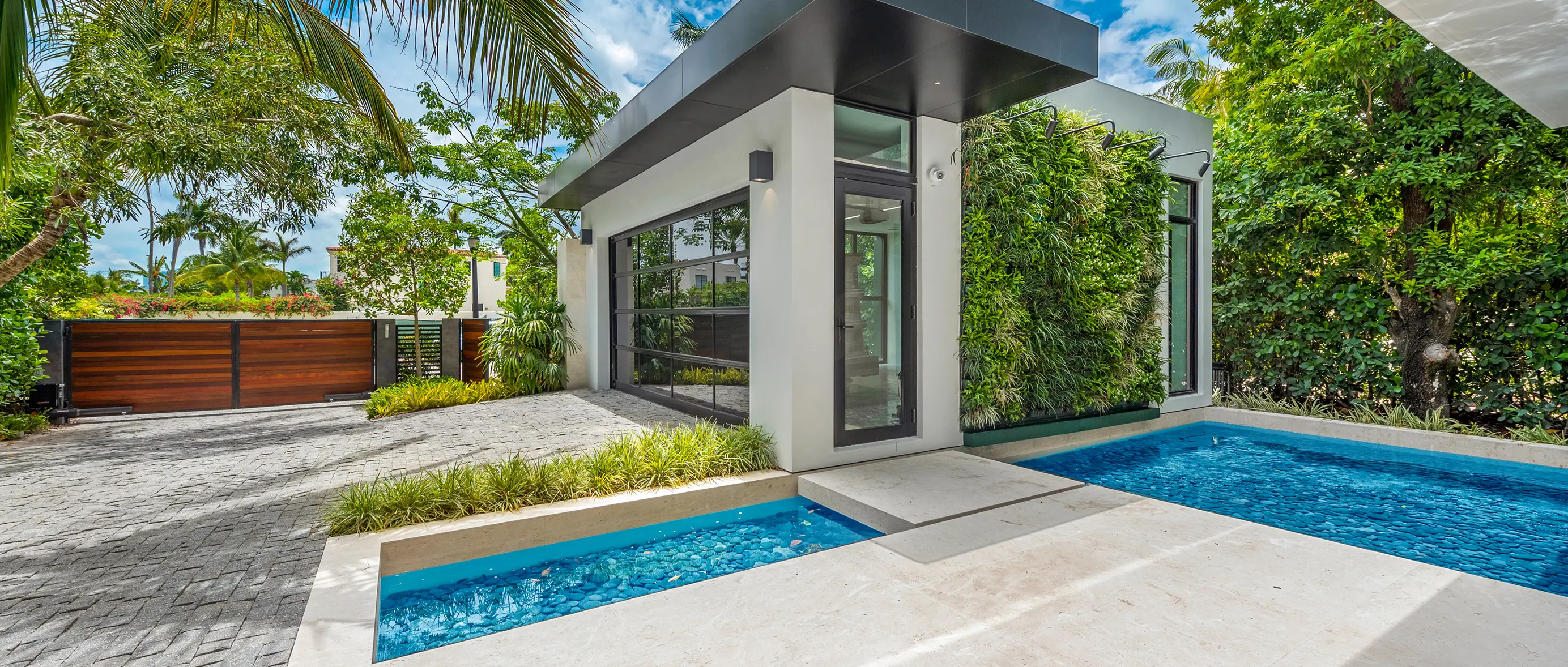 Front driveway with modern architectural entrance, infinity pool moat, and lush tropical landscaping under blue sky