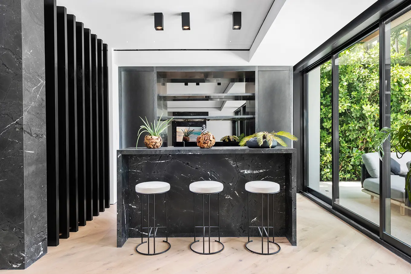 Contemporary bar area with black marble island, white leather stools, and floor-to-ceiling windows overlooking tropical garden