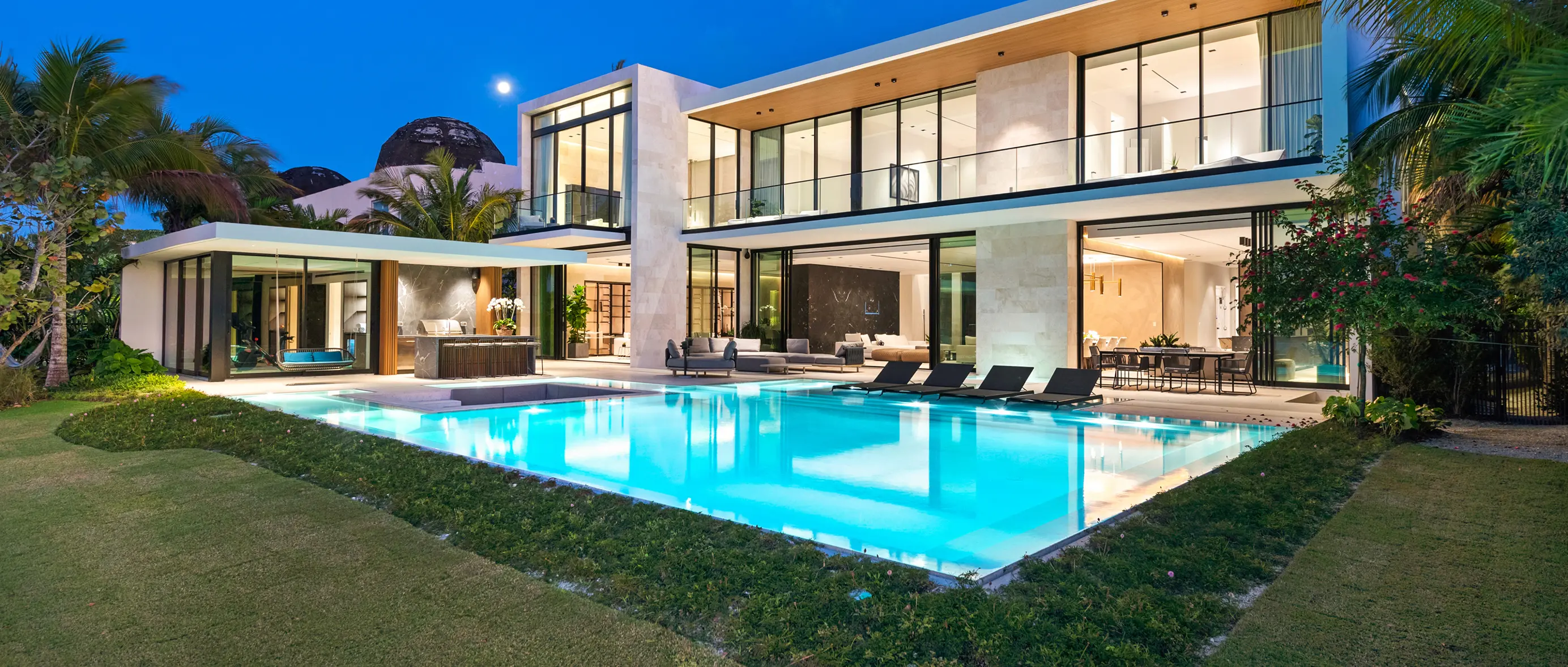 Contemporary two-story estate illuminated at twilight with glowing infinity pool, floor-to-ceiling glass walls, and lush tropical landscaping