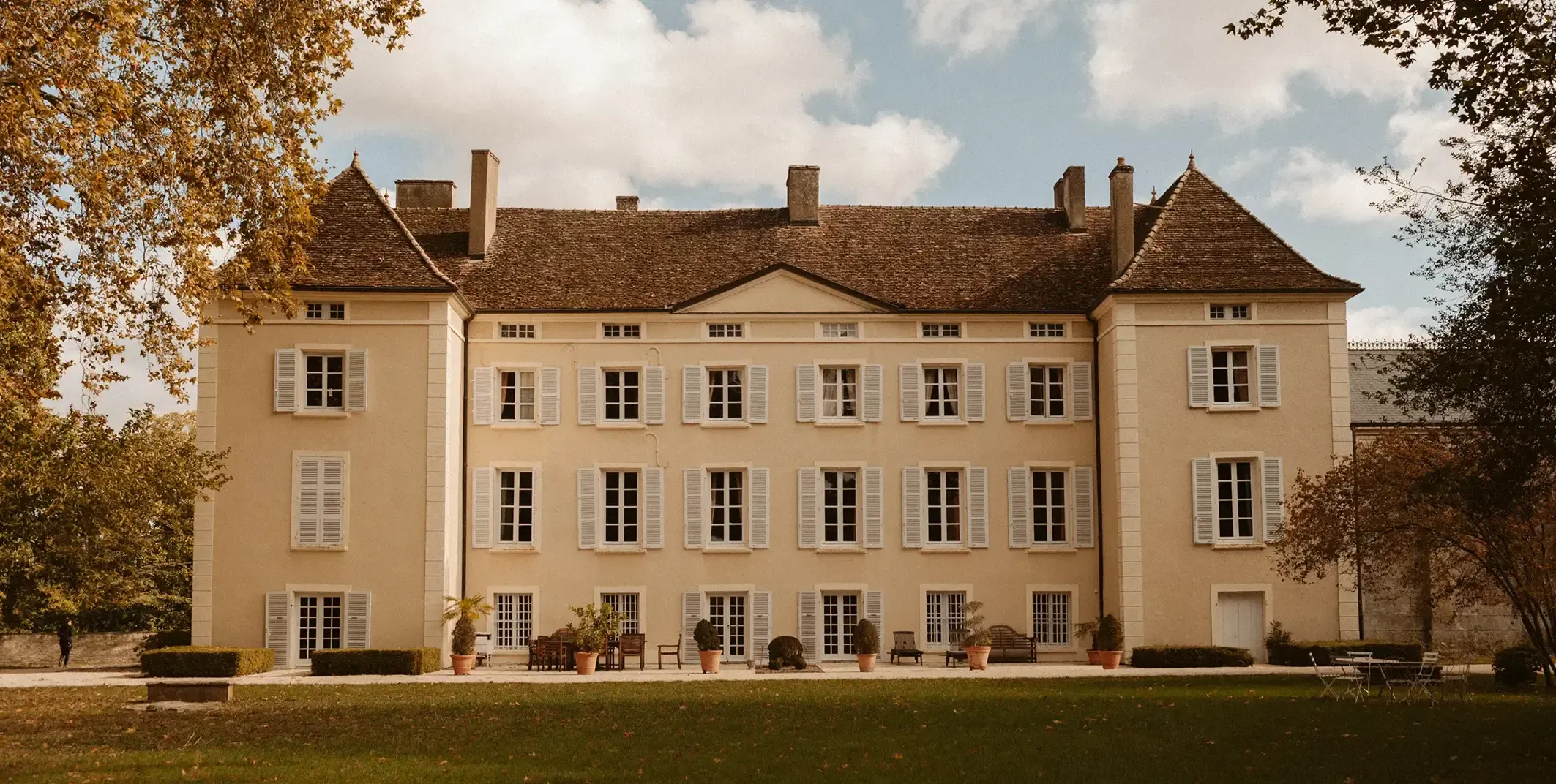 Façade ensoleillée d'un château bourguignon aux volets blancs, entouré d'un jardin avec terrasse.