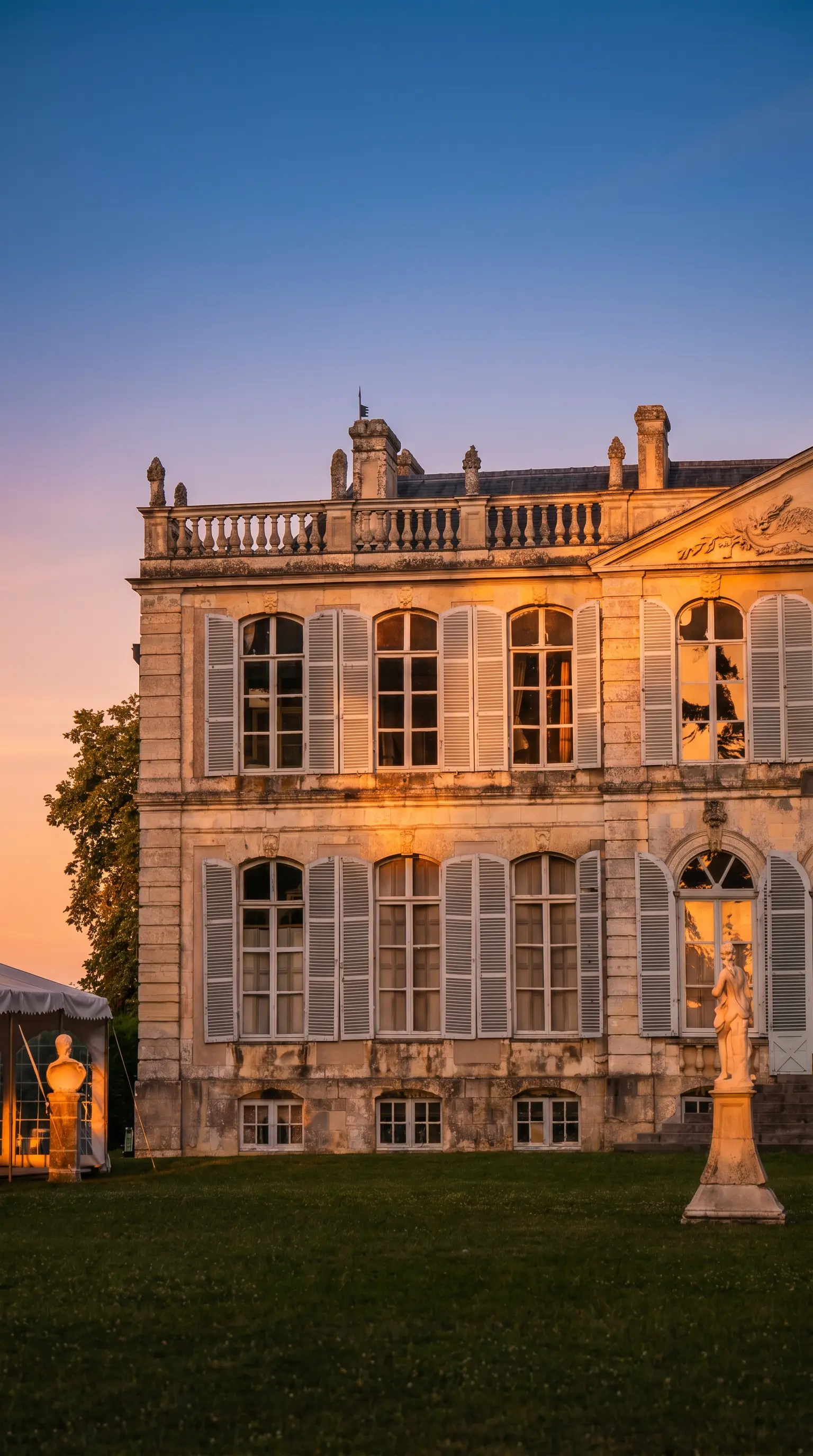 Le château de Canon en pierre avec des volets bleus et des détails architecturaux ornés se dresse sur une pelouse herbeuse à côté d'une statue blanche et d'une structure de tente.