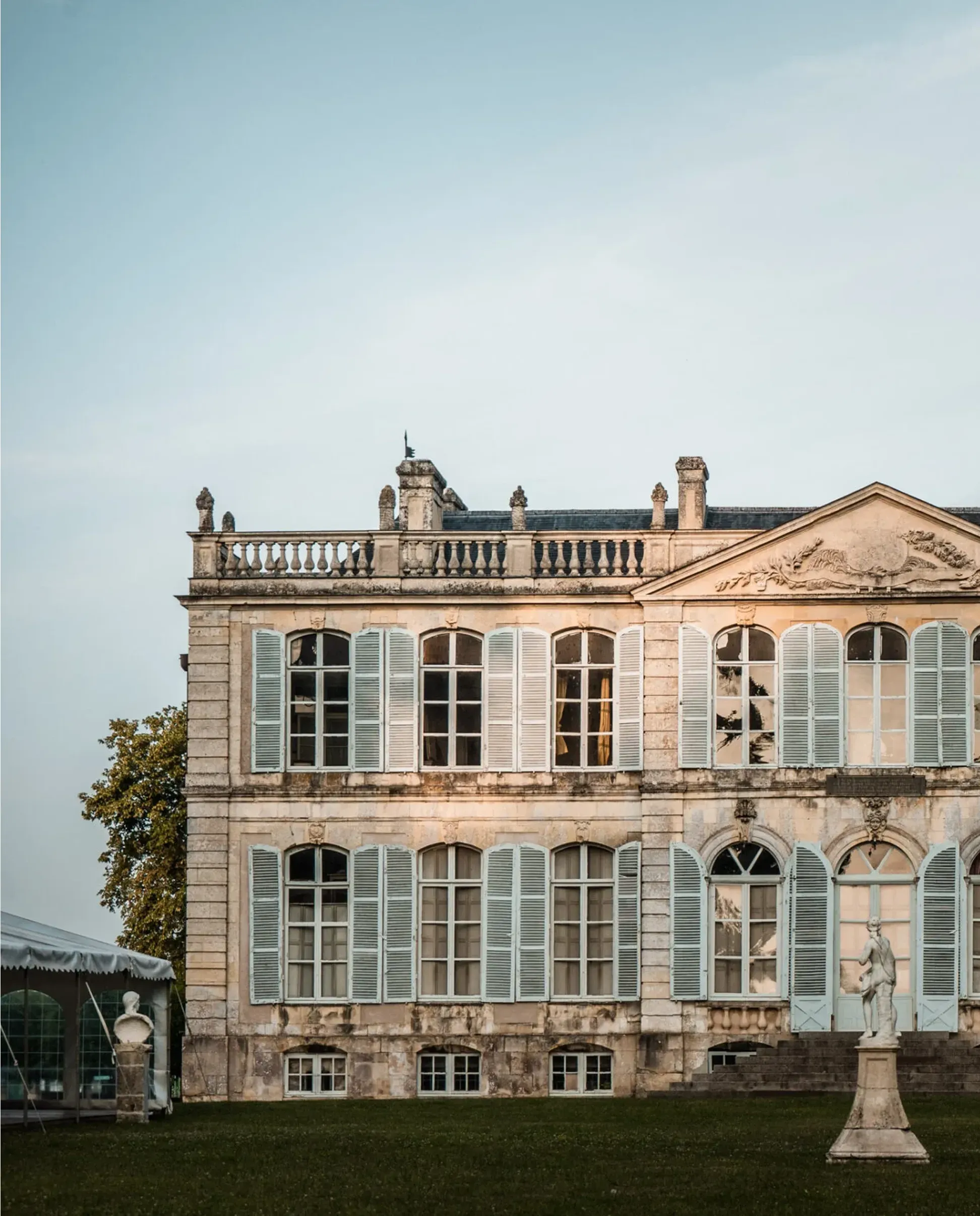 Canon's stone castle with blue shutters and ornate architectural details stands on a grassy lawn next to a white statue and tent structure.