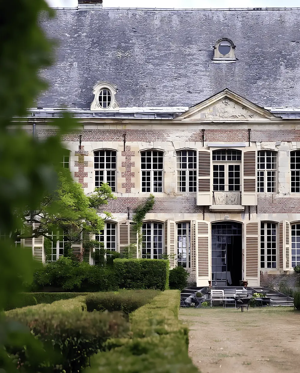 European-style Château de Bernoville with high windows, open shutters and manicured hedges in the foreground, seen on a cloudy day.
