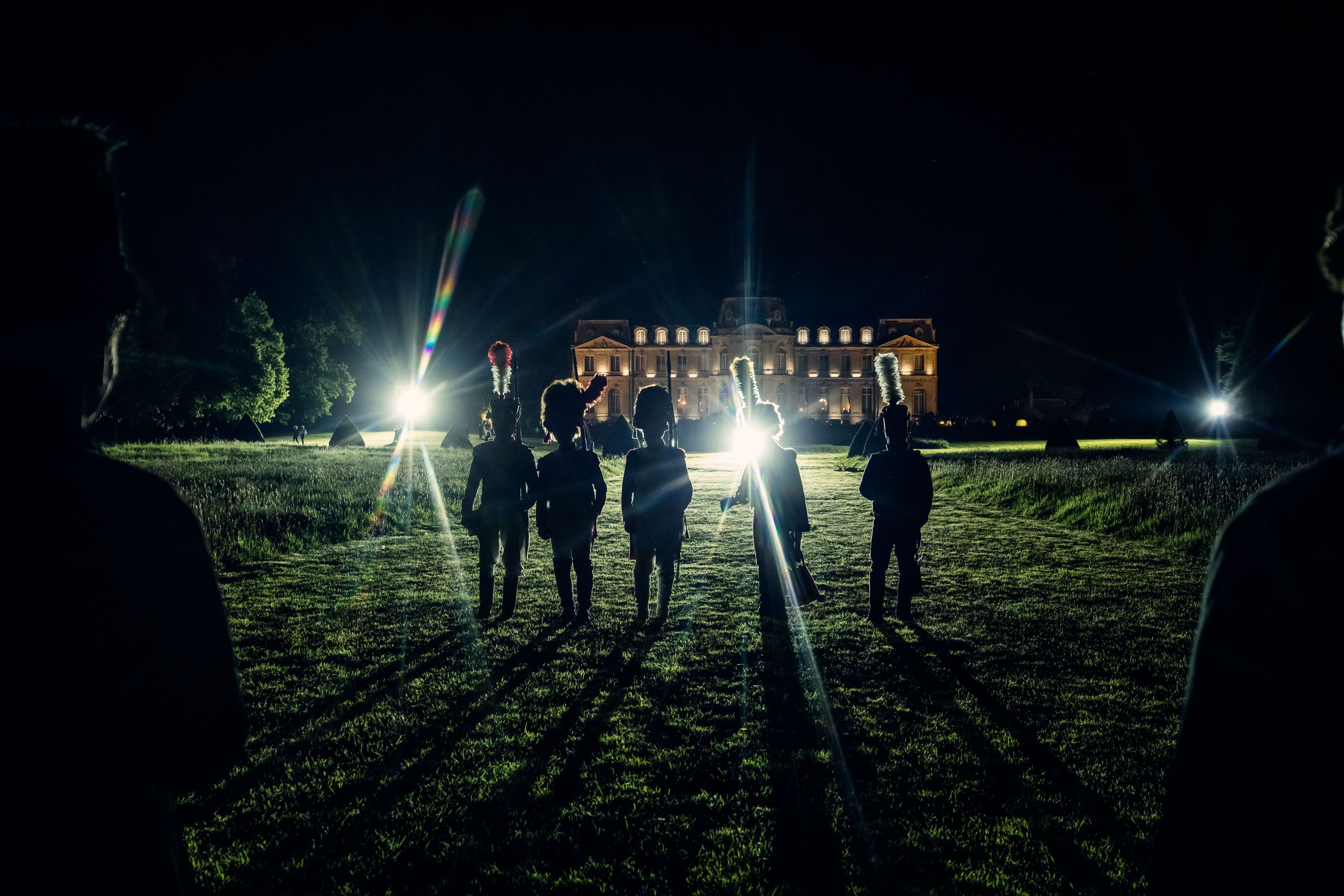 Cinq silhouettes en uniformes sombres se tiennent de dos sur un chemin herbeux la nuit, face à un château fortement illuminé avec un contraste marqué.