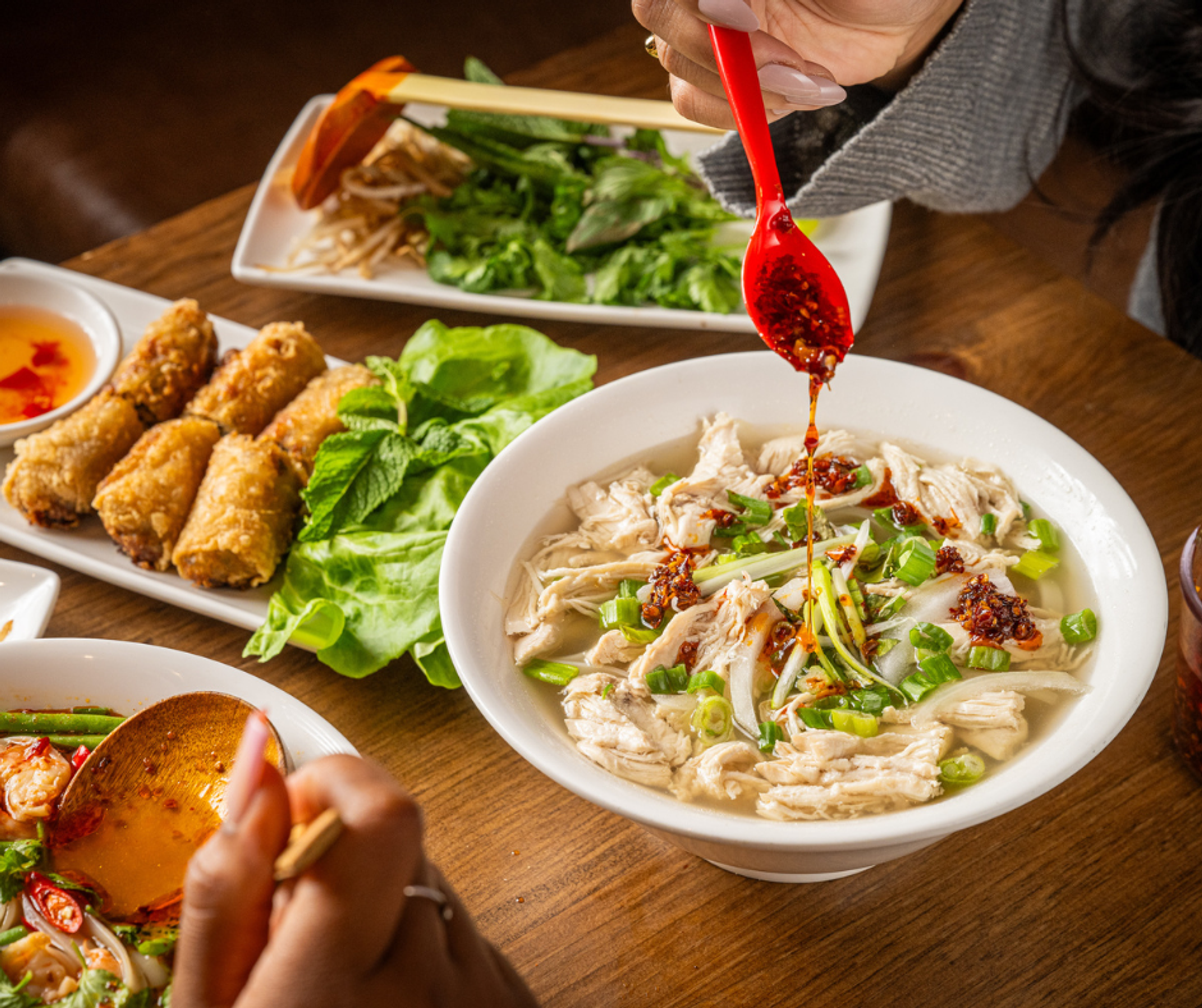 A woman's hand drizzing chili oils into a bown of chicken soup, with other food dishes on the table in the background.