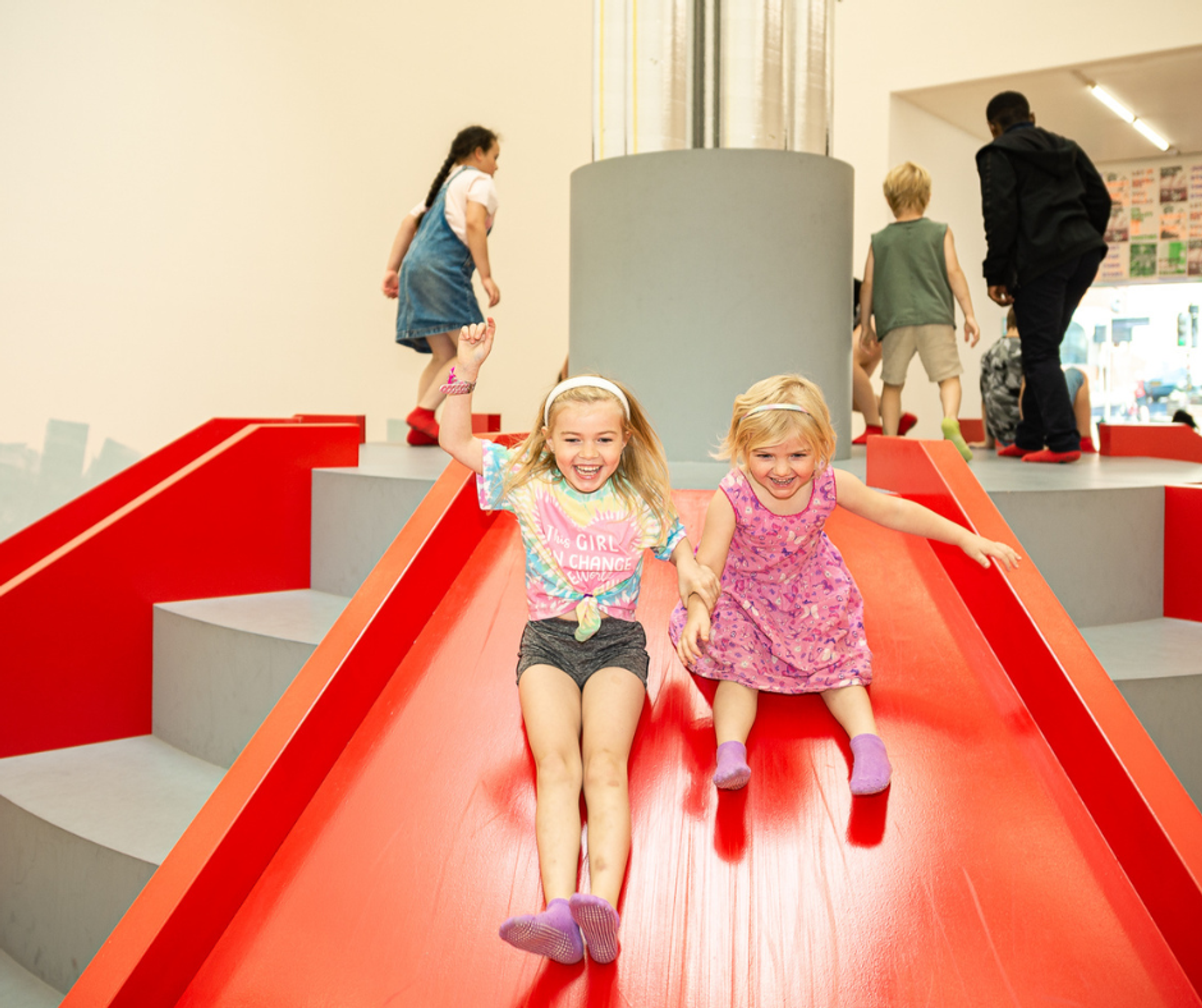 Two young girls laughing as they go down a slide in an indoor play area