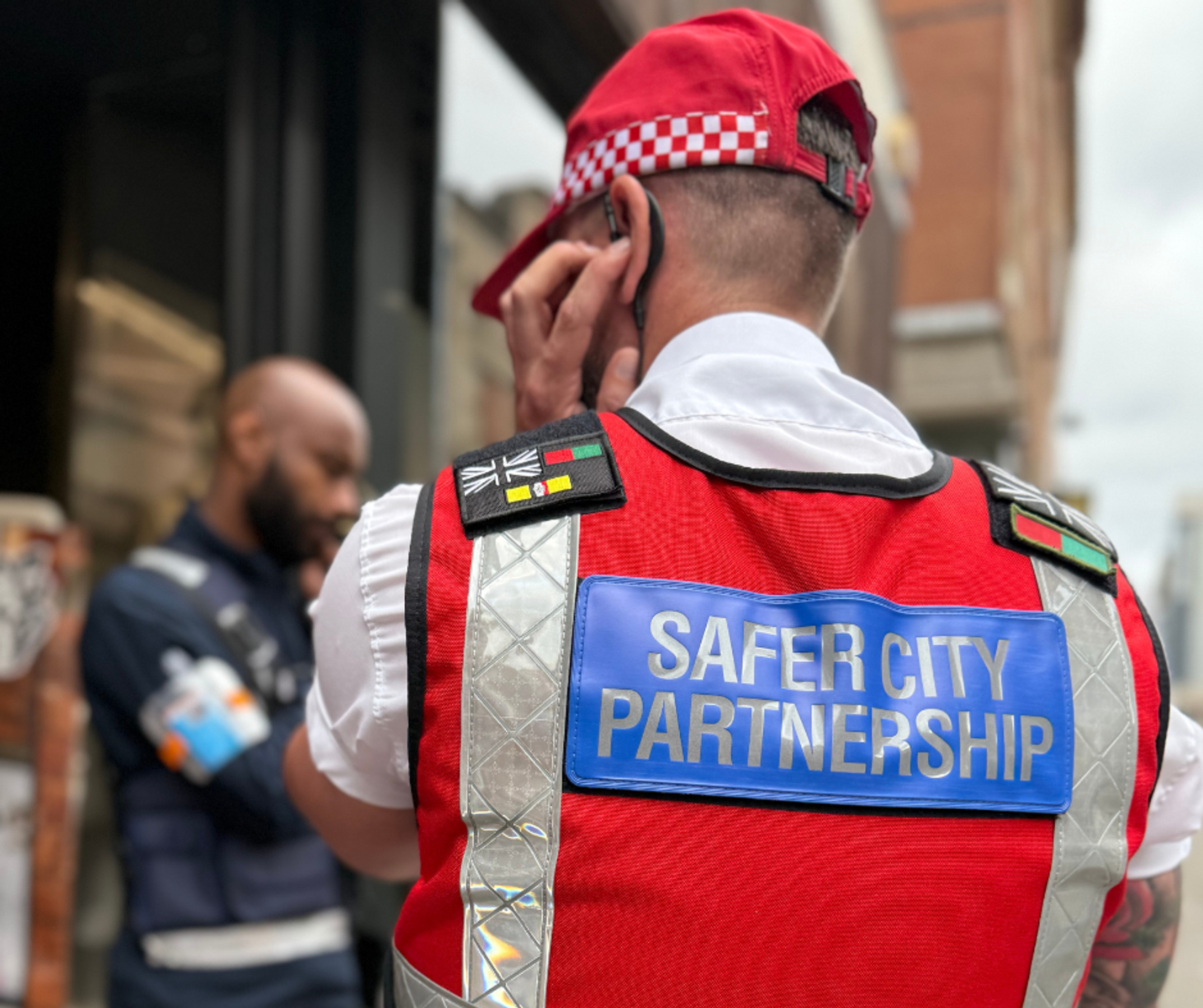 The back of a man in a red tactical vest which reads 'Safer City Partnership'. He is wearing a red cap and holding an earpiece to his ear.