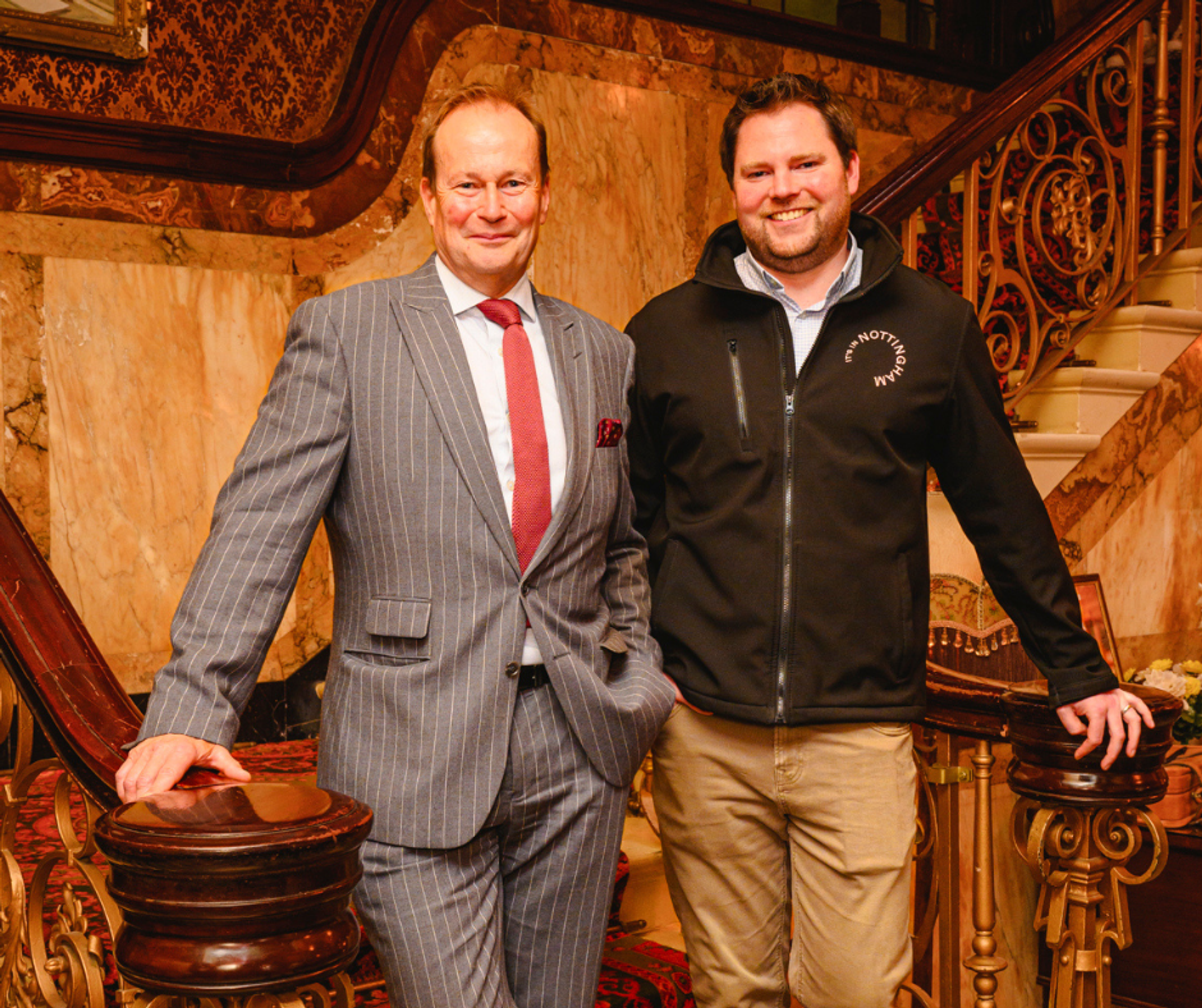 Two men stood at the foot of an ornate stairwell smiling at the camera