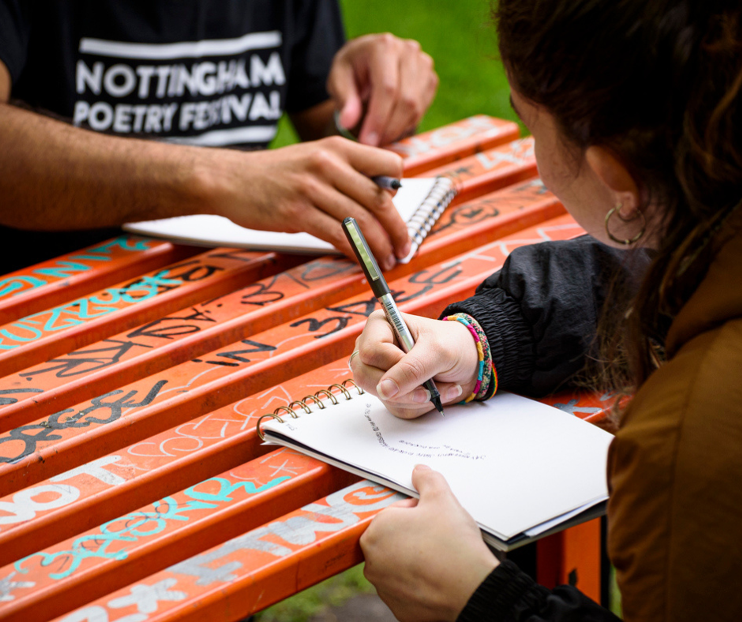 A woman and man sketching ideas in their notebooks sat at a graffitied park bench.