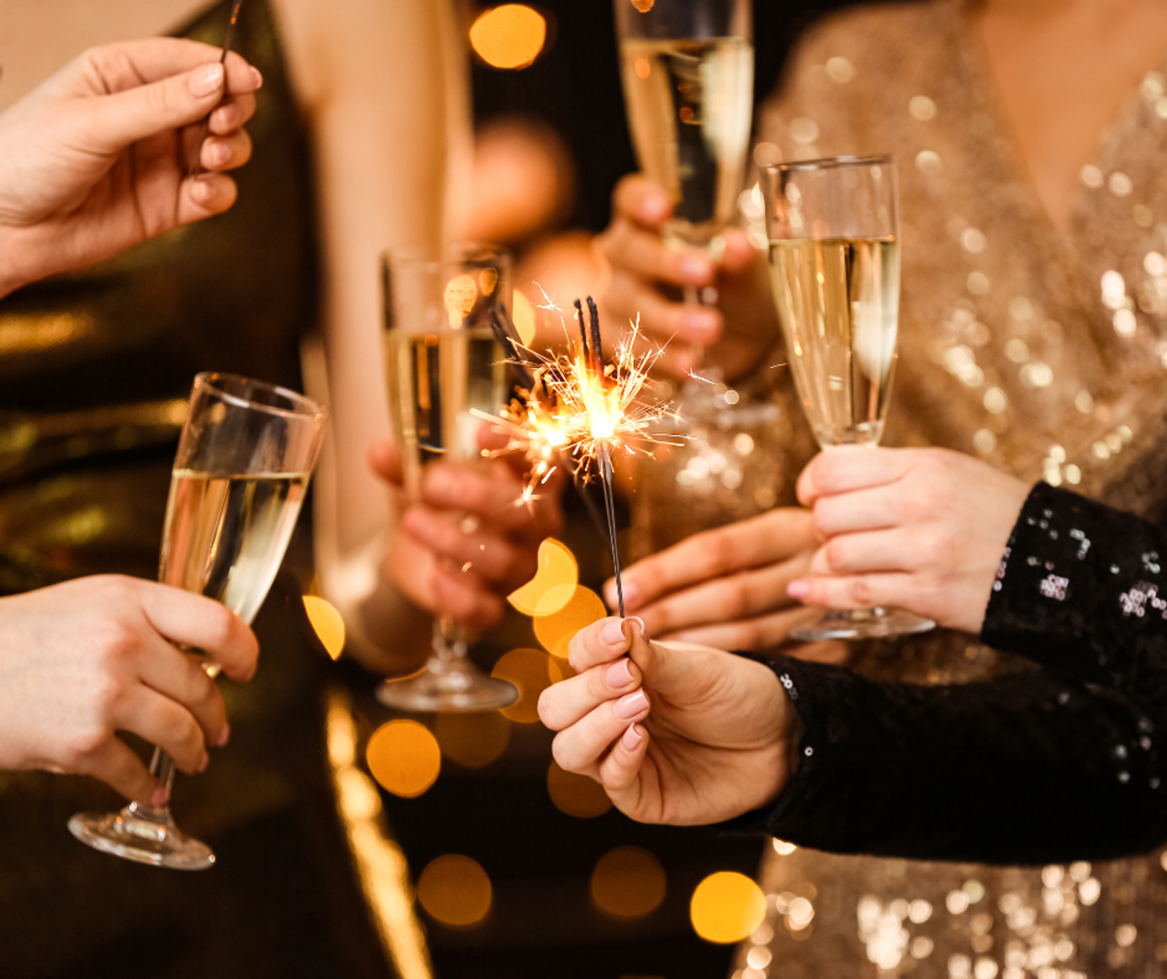 4 hands holding glasses of champagne to toast a celebration, with two hands holding in sparklers. The people are wearing gold and black sparkly outfits.
