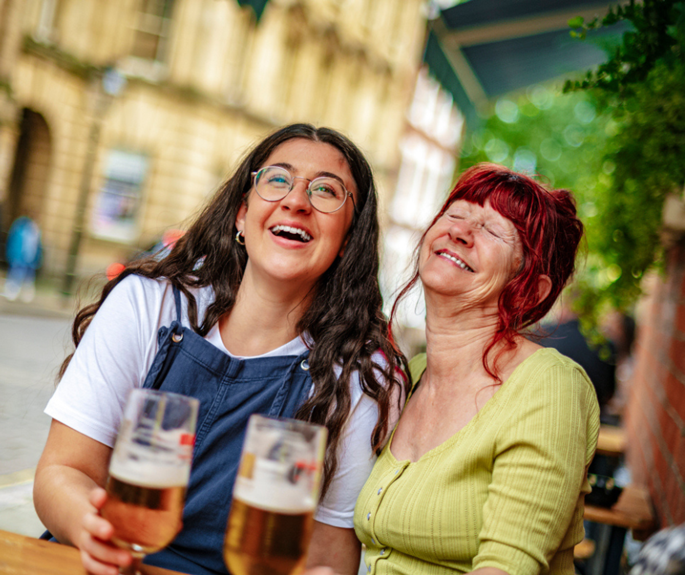 A mother and daughter sat laughing and smiling at a table outside a bar with beers