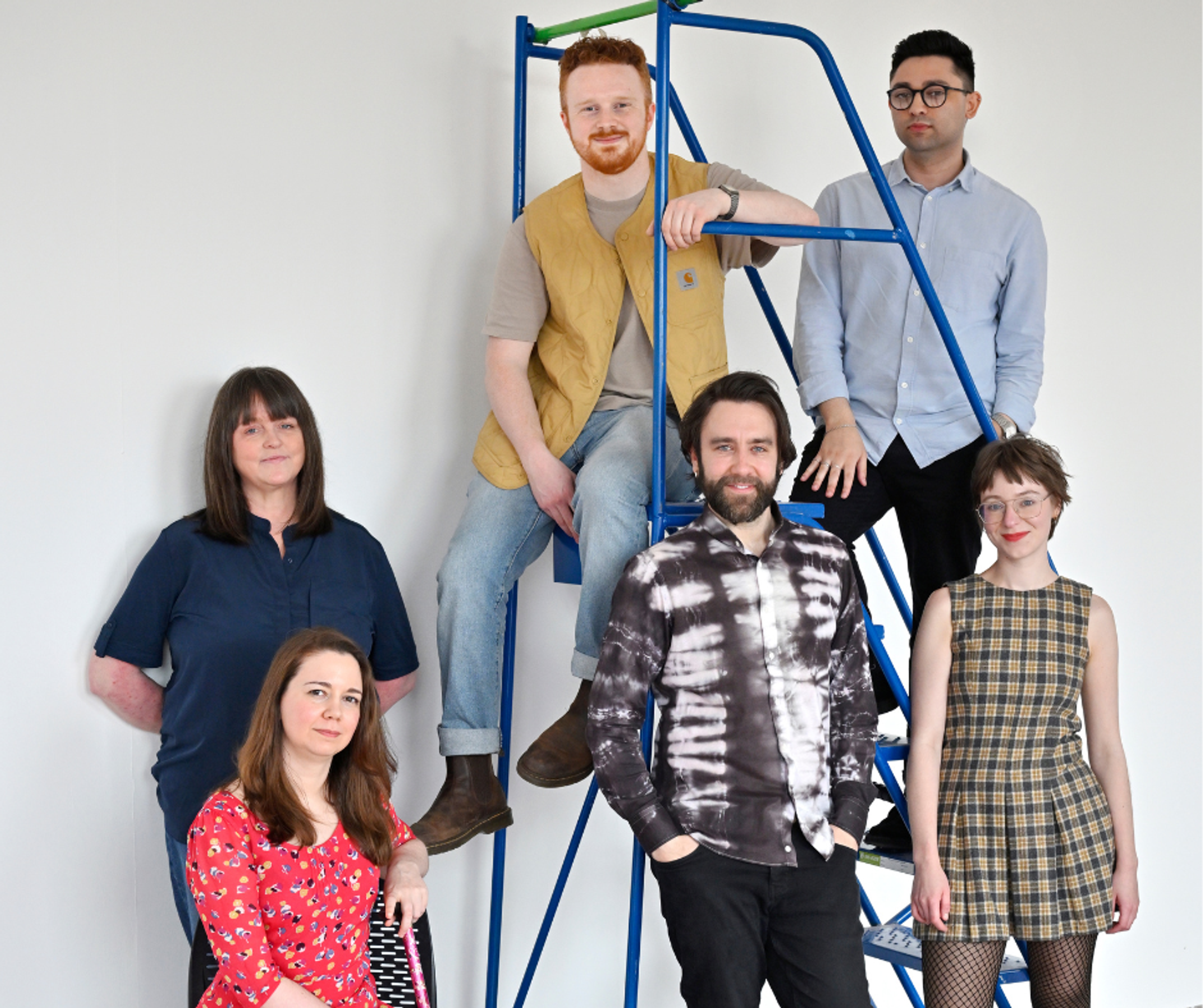 A group of people against a white wall, with two of them standing and sitting on a large blue step ladder.
