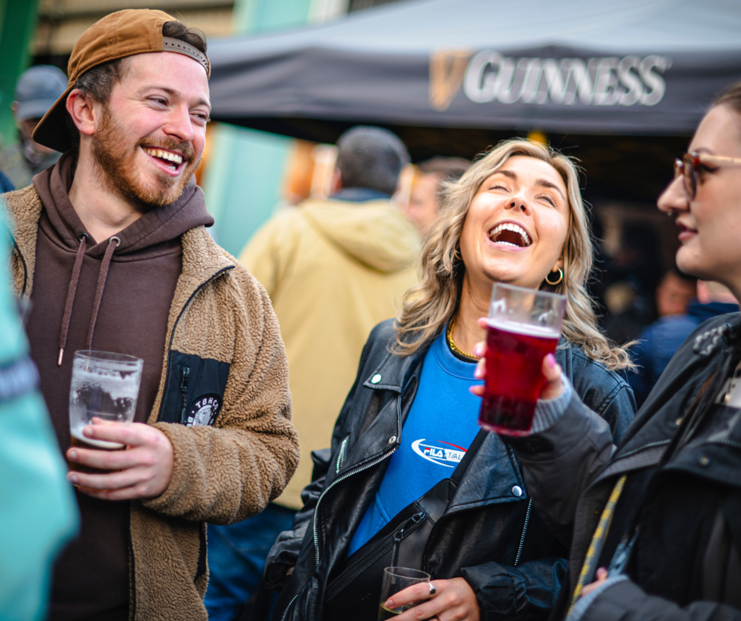 A man and two women outside laughing. They're holding beer and there's a beer tent visible in the background.
