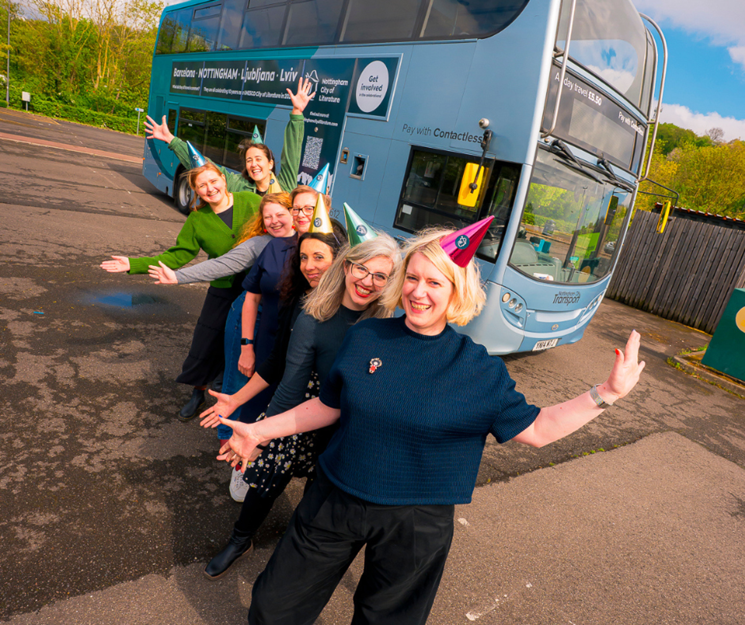 A row of women wearing party hats, smiling and posing with their arms up in front of a Nottingham bus.