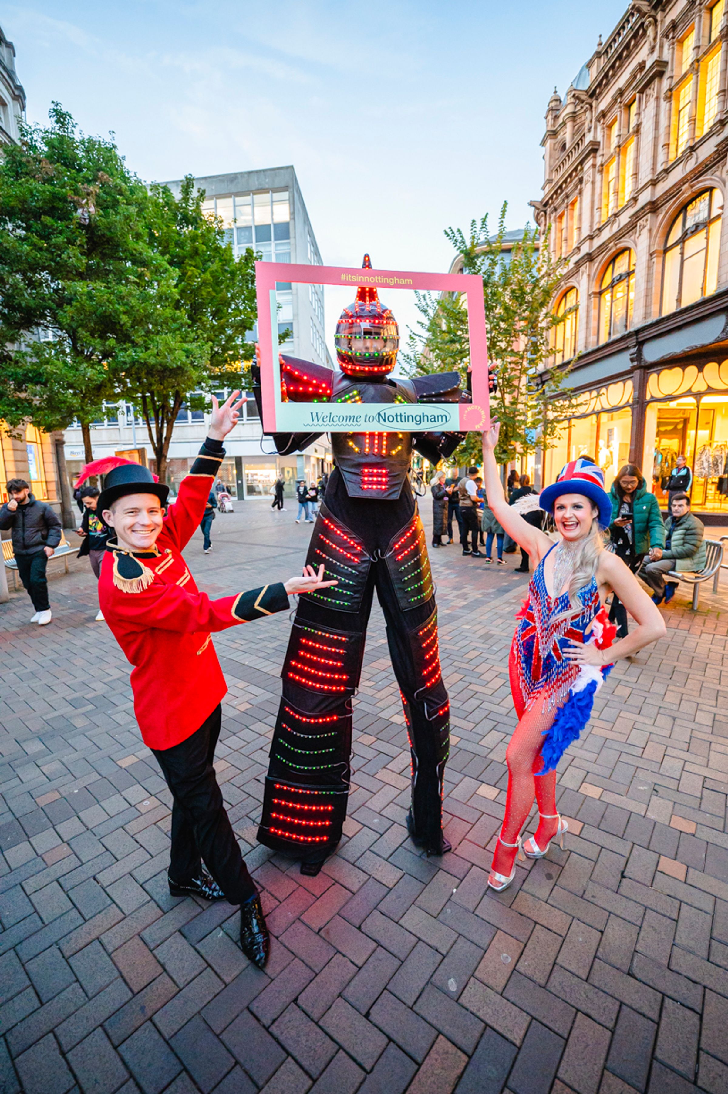 Street entertainers in Nottingham in bright costumes, one is in stilts and holding a frame that says "Welcome to Nottingham"