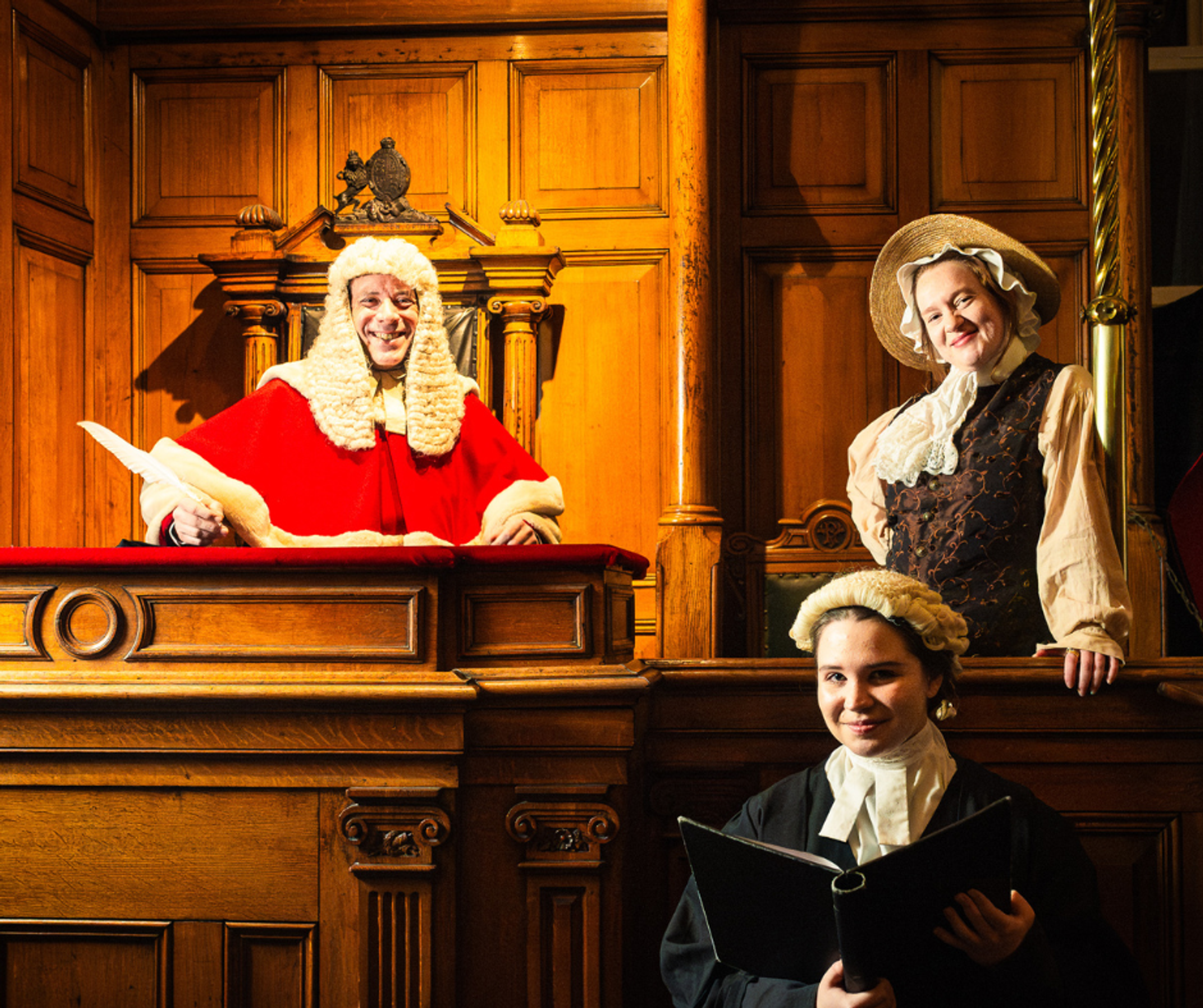 Cast members at the National Justice Museum - a judge smiling in a red cape and wig and lit up by a spotlight, a woman smiling from the witness stand and a young girl in wig holding a folder and smiling.