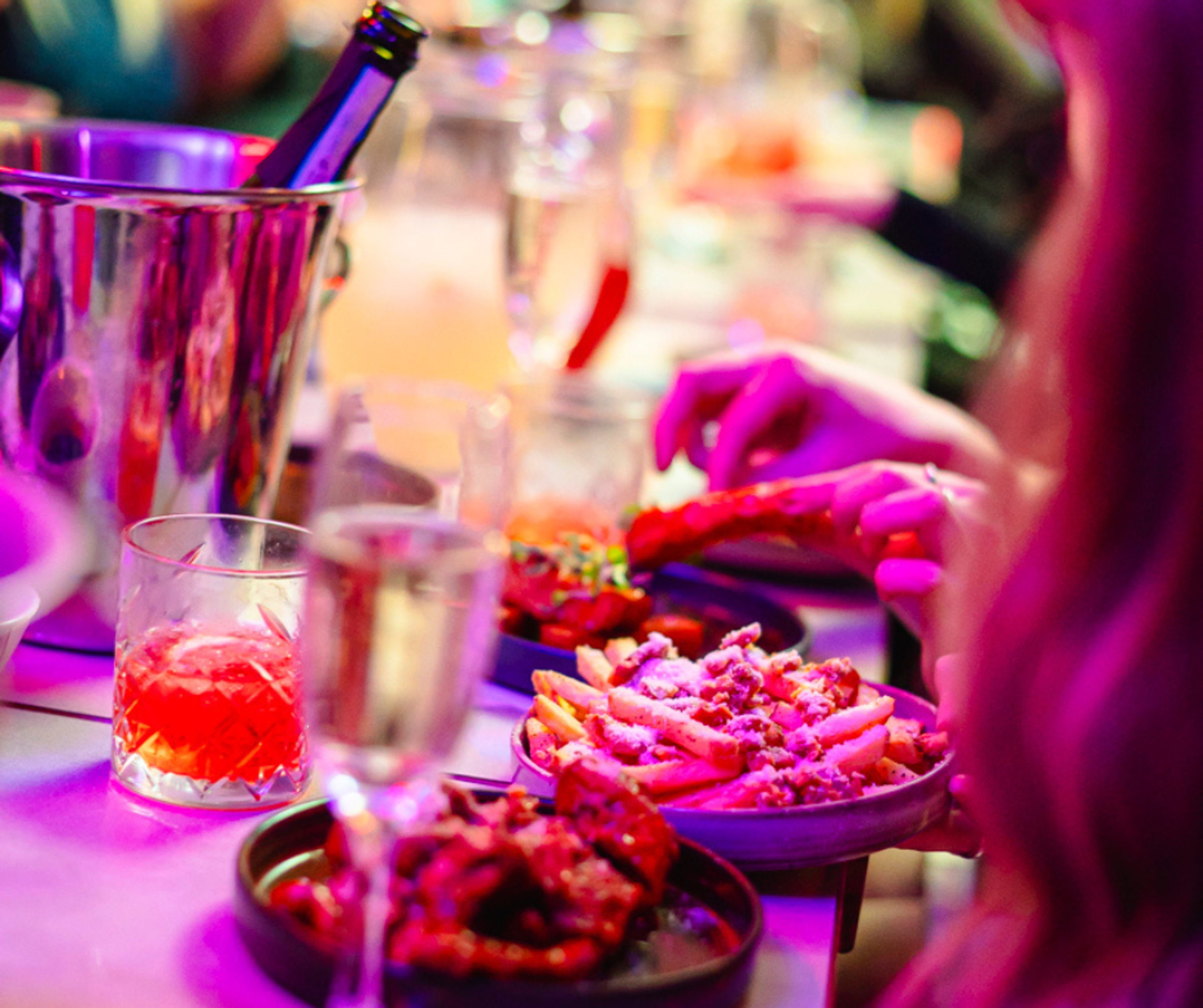 An over-the-shoulder shot of bottomless brunch, including a few plats of food, cocktails and a bottle of bubbles on ice.