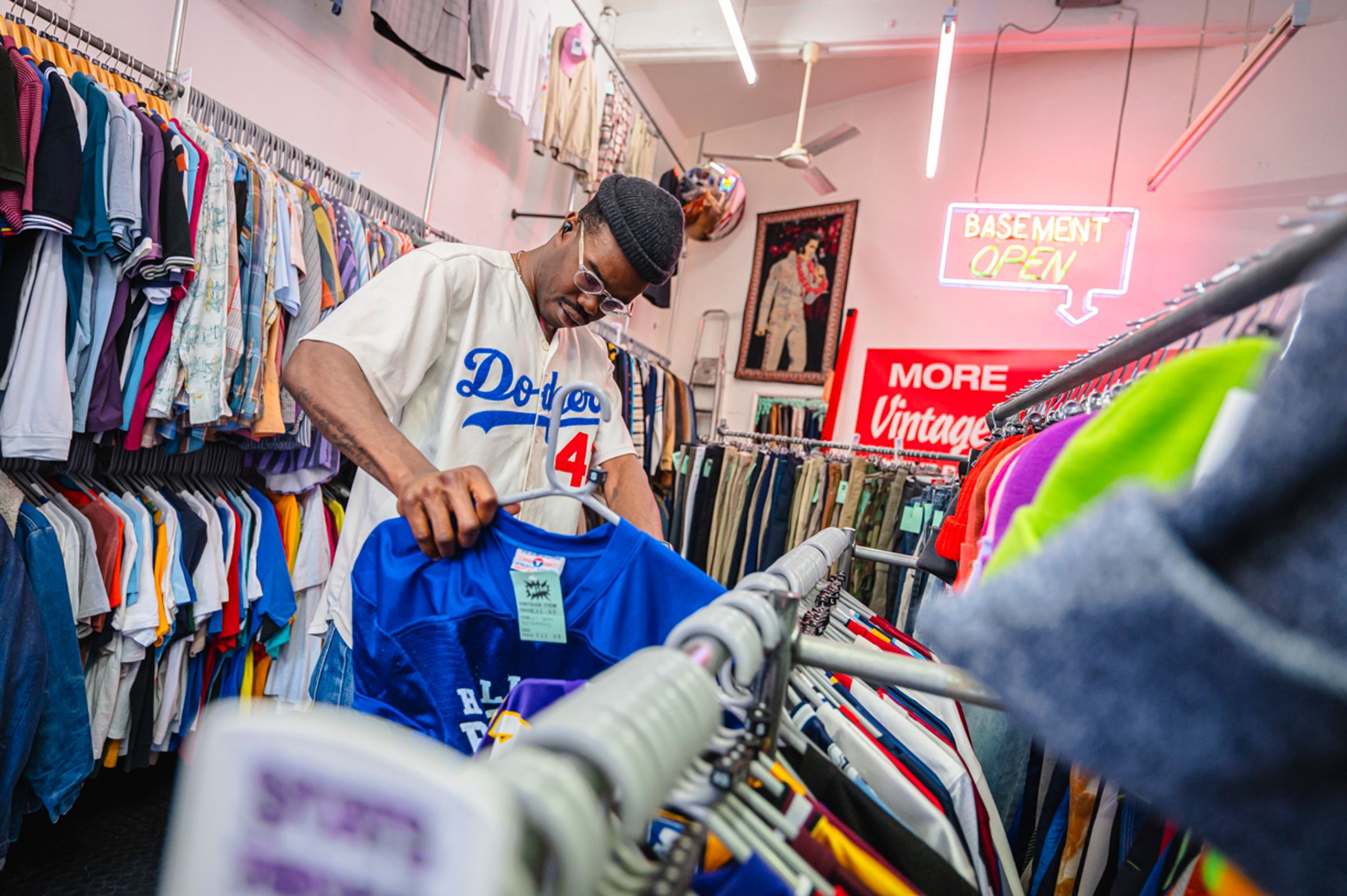 A man browsing racks of vintage clothing in a full shop. A sign in the background reads "basement open"