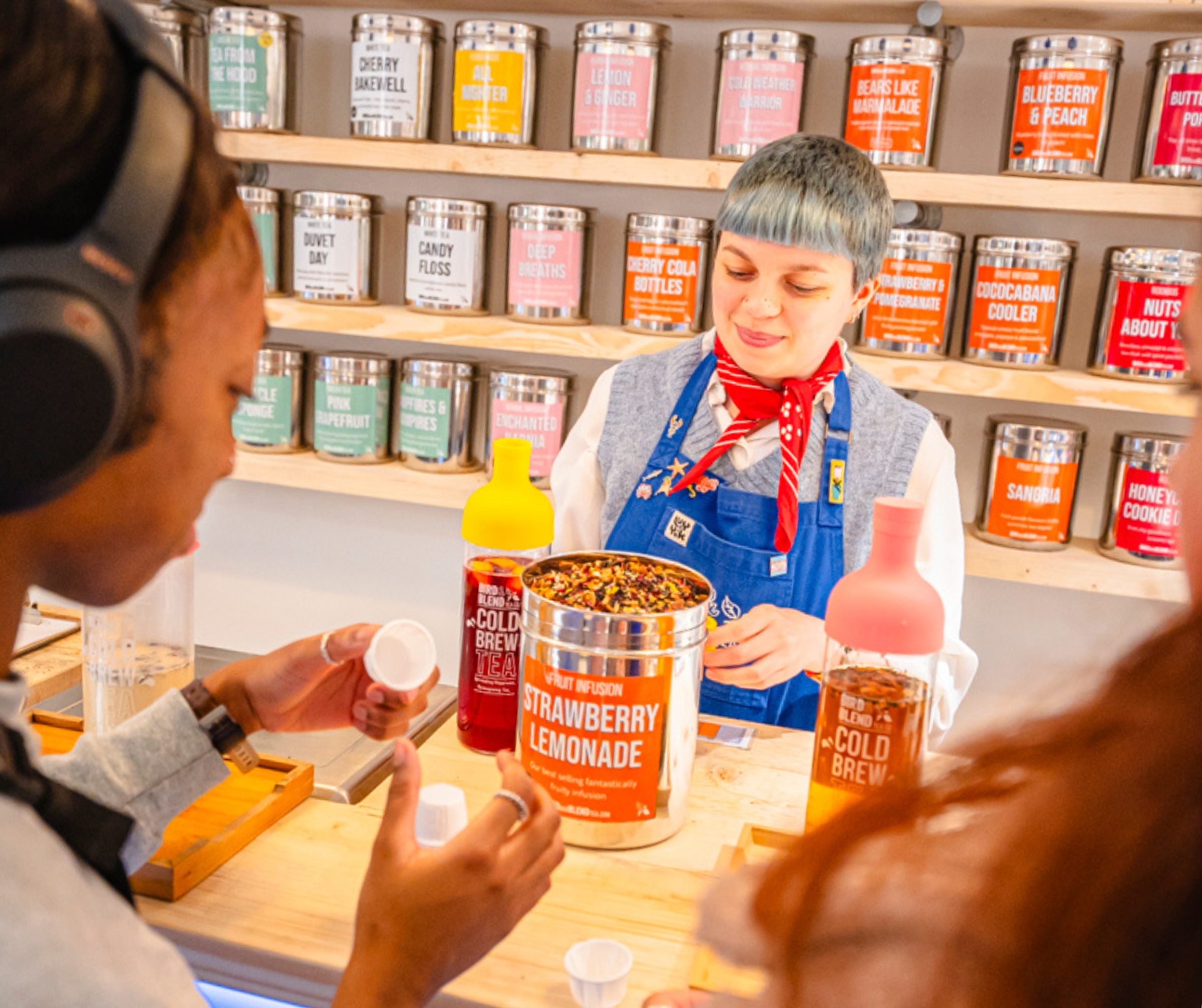 A shop assistant in a blue apron giving tasters of a tea from a tin labelled 'strawberry lemonade'
