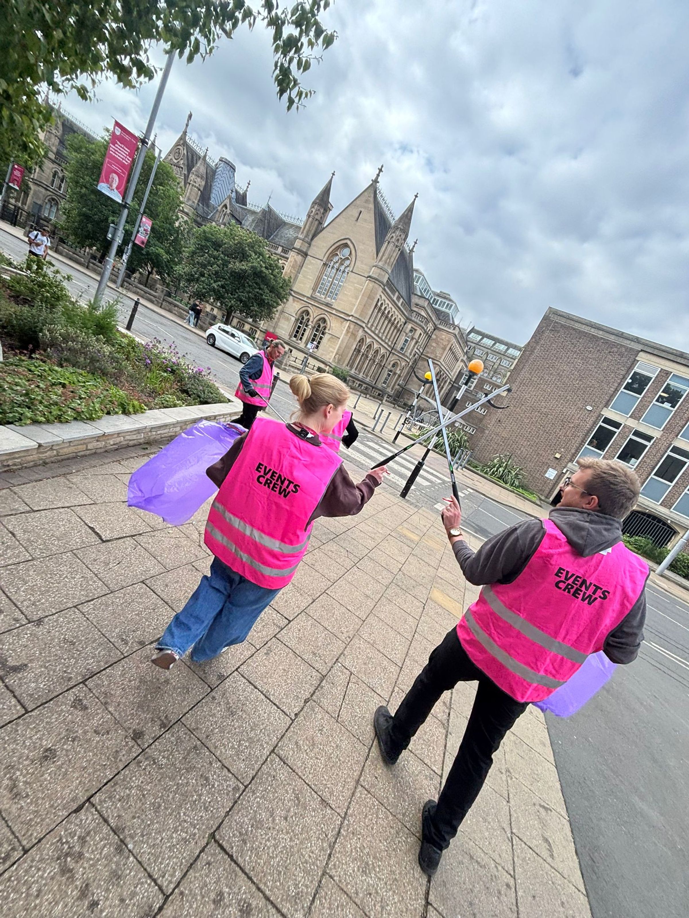 Walking along behind two litter pickers on the Nottingham Trent University campus. They are wearing pink high vis vets and jokingly jousting with their litter pickers.