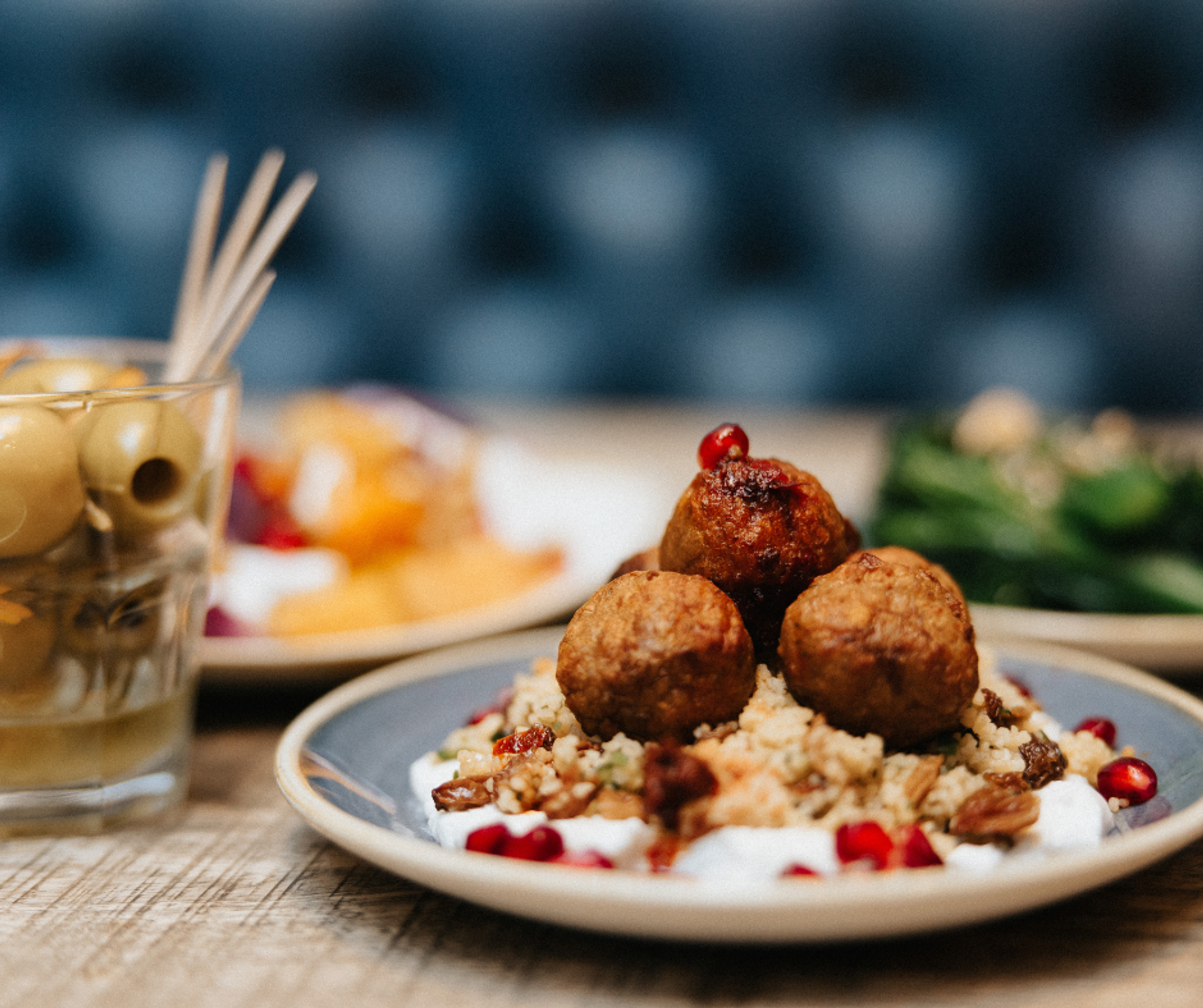 A plate of meatballs on couscous with pomegranate seeds and yoghurt, next to a glass of olives as part of a restaurant spread.