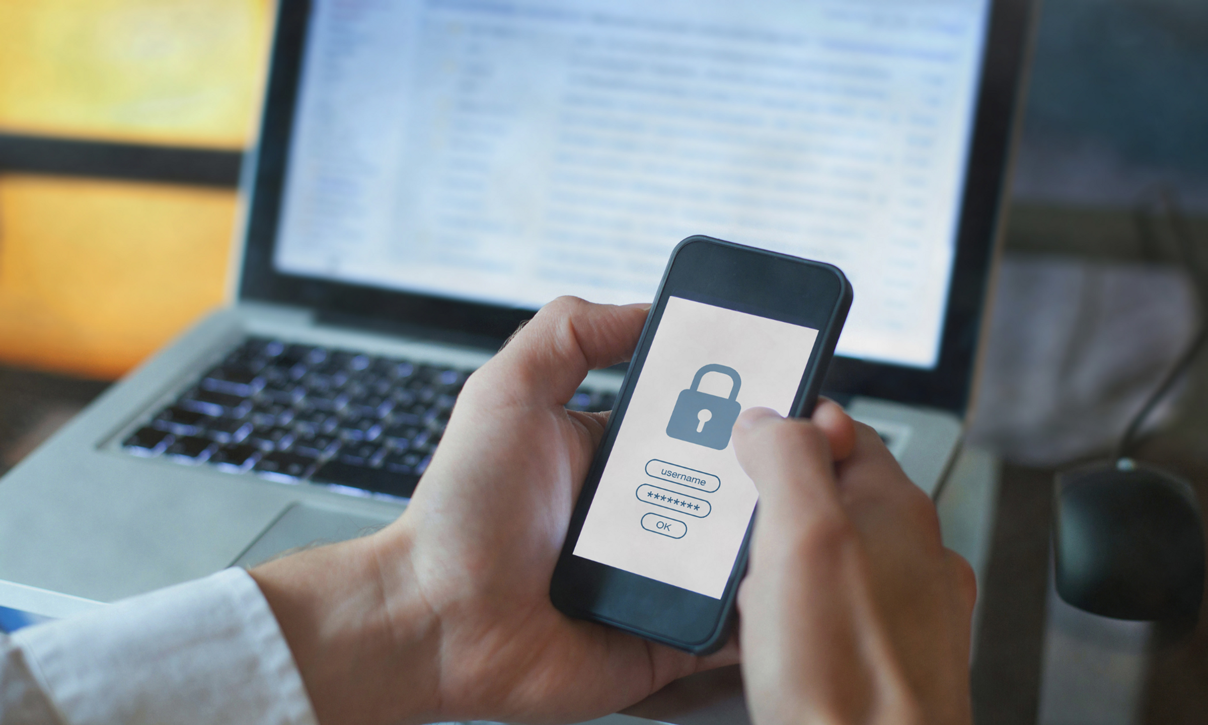A man holding a mobile phone with a security padlock screen on it, with a laptop in the background.