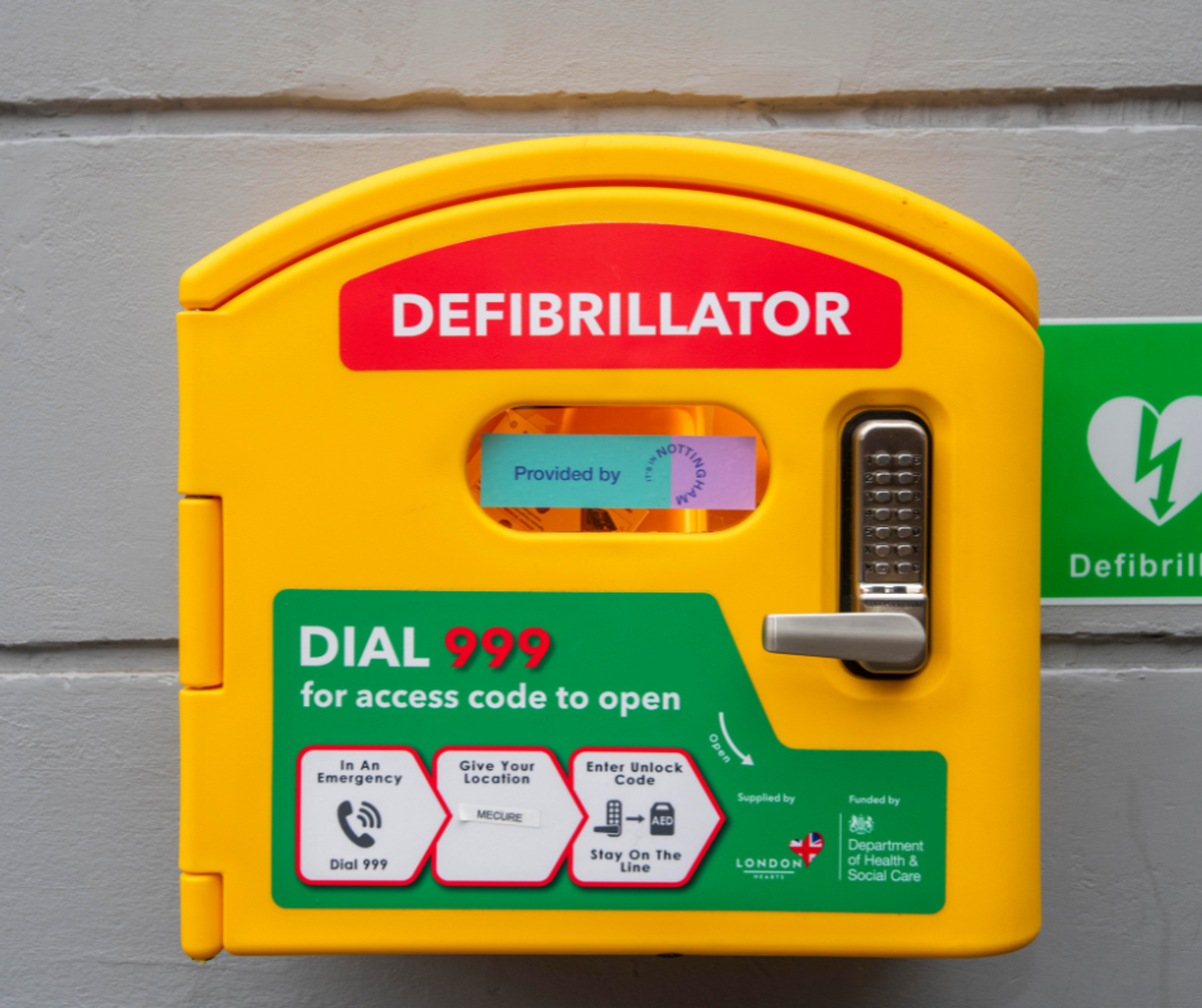 A yellow defibrillator case with a sticker reading "provided by It's in Nottingham". Installed on a grey wall.