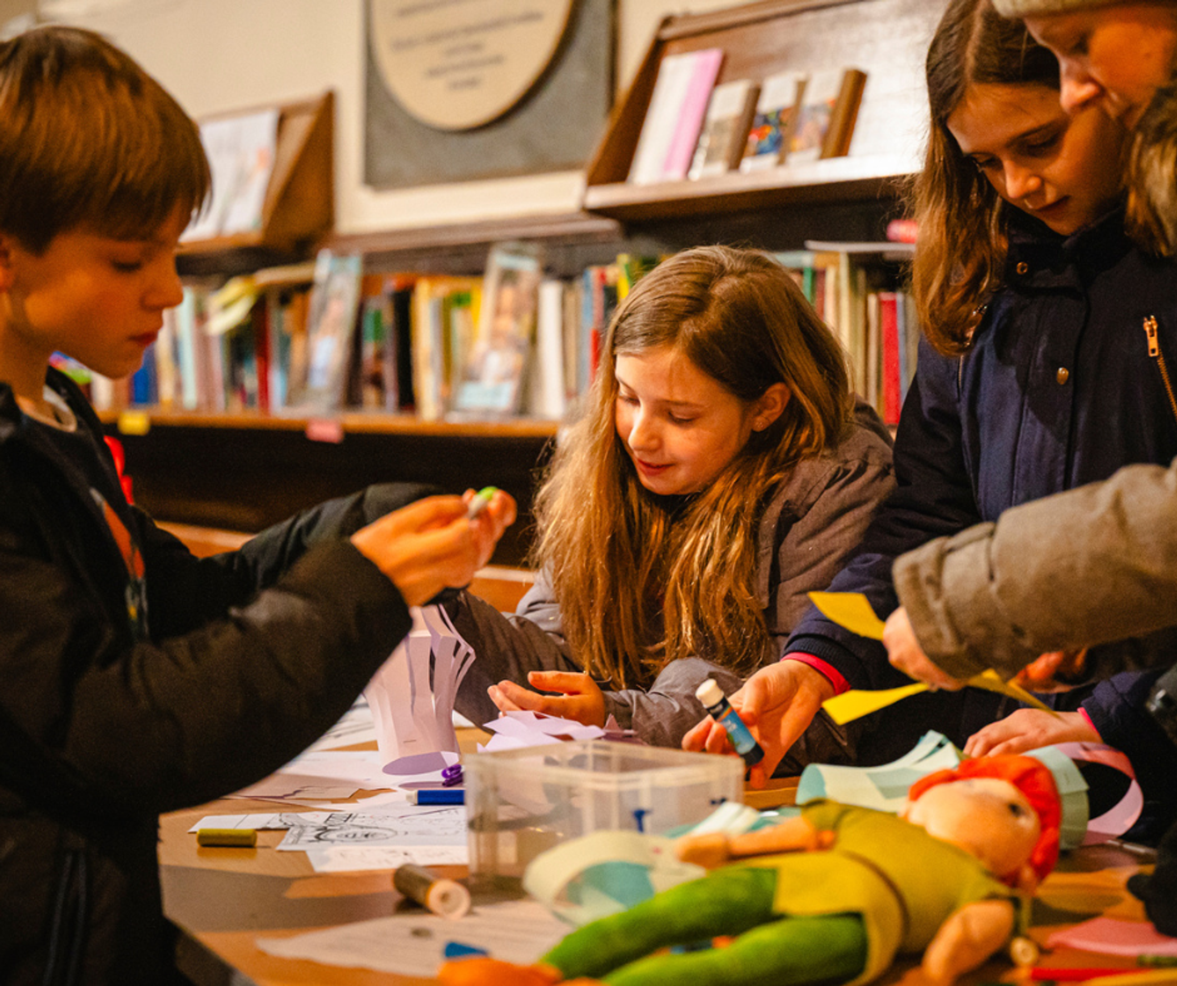 A group of children around a table making paper lanterns. They're wearing outdoor winter coats.