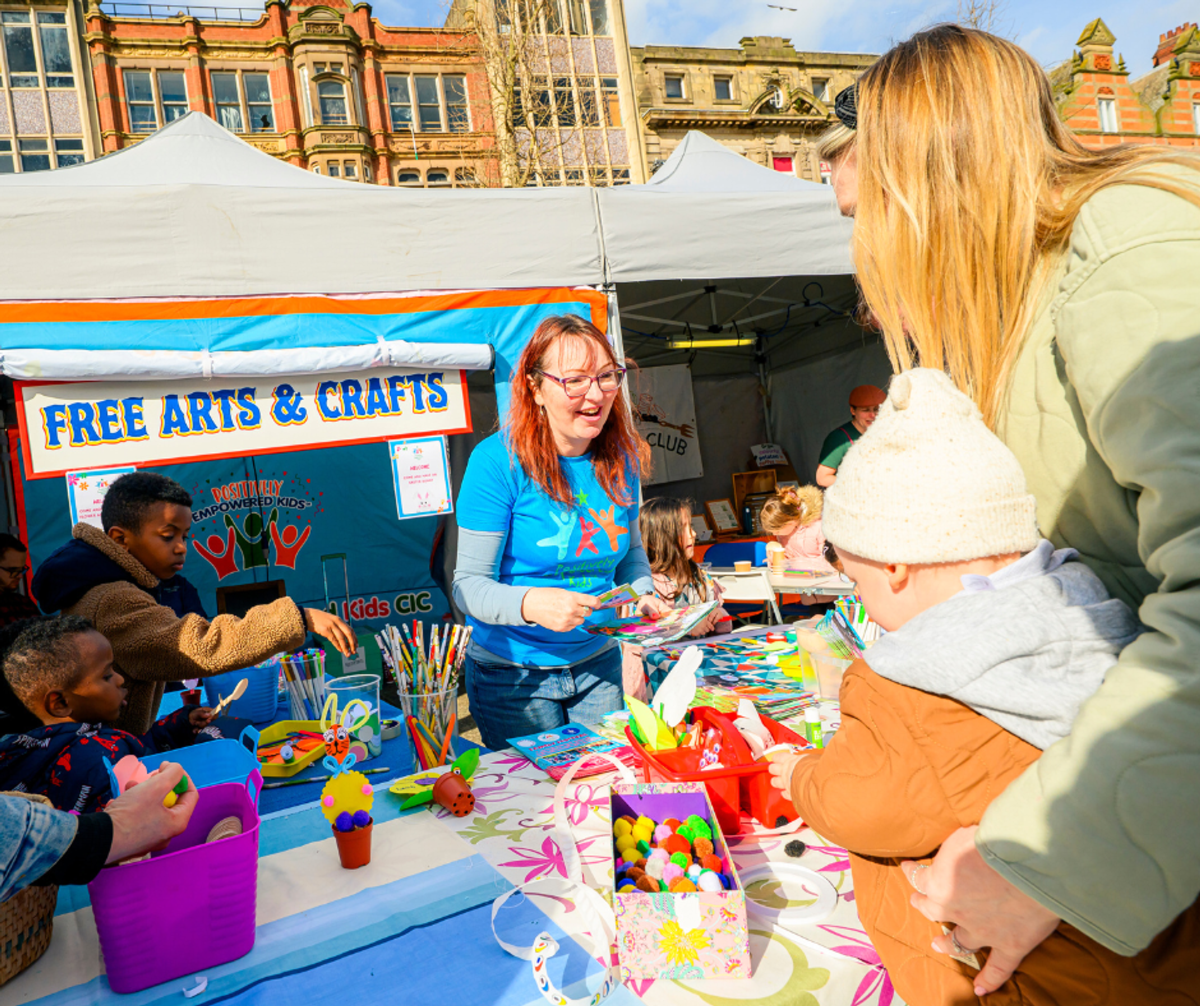 A smiling activity provider at an outdoor market as a parent and child look at the arts and crafts activities on offer.