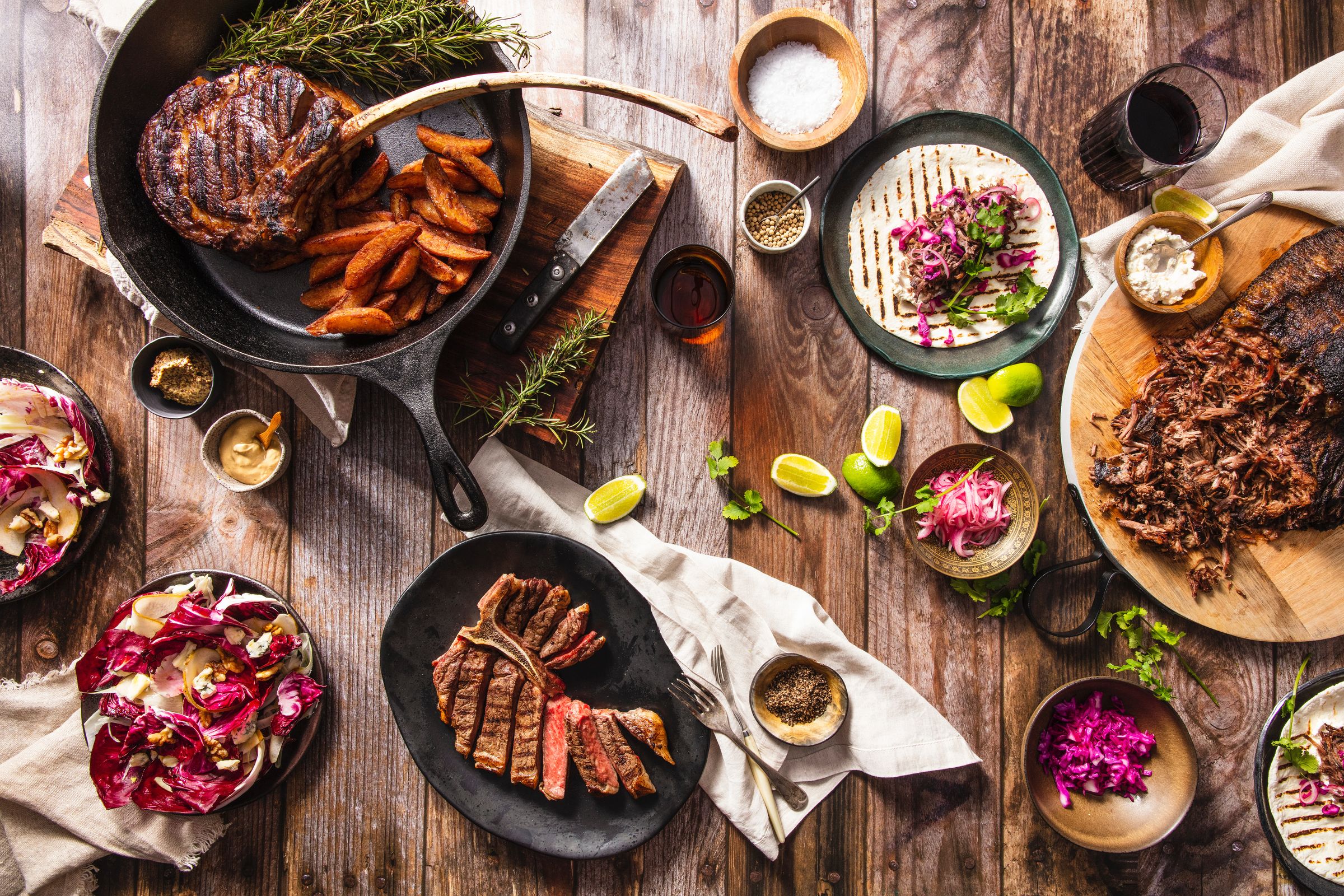 An overhead shot of a table with plates of steak, lamb and other meat with side dishes and garnishes