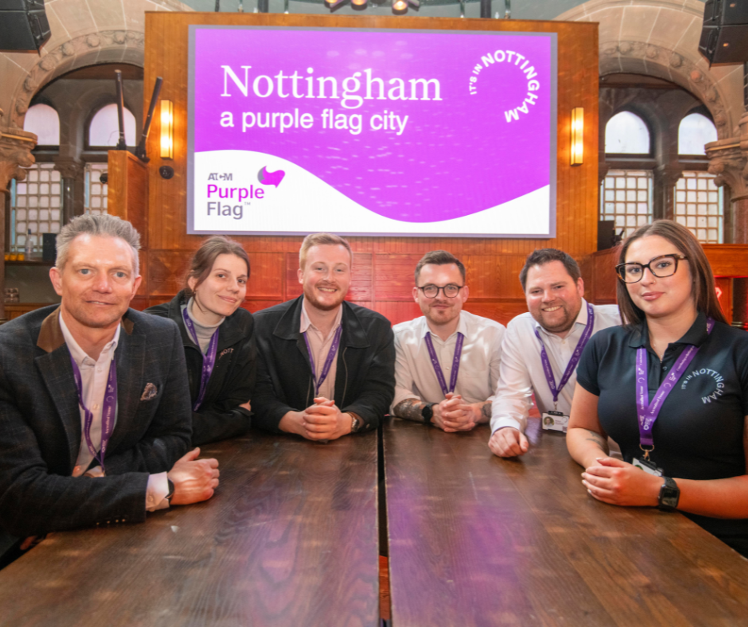 Members of the It's in Nottingham team sat round a table facing the camera, with a projection in the background reading "Nottingham: a purple flag city"