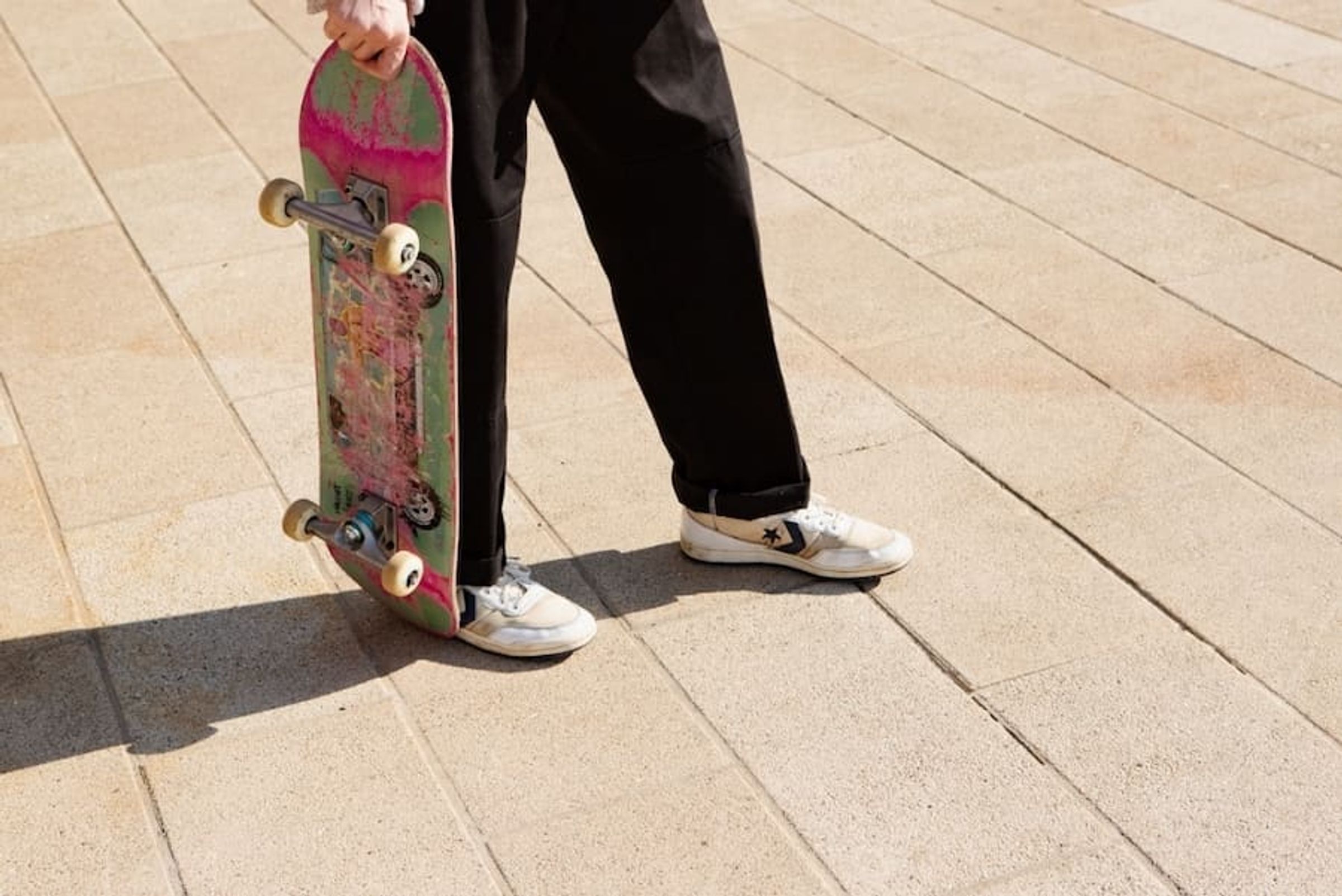 The legs and hand of a skateboarder in black trousers holding their skateboard.