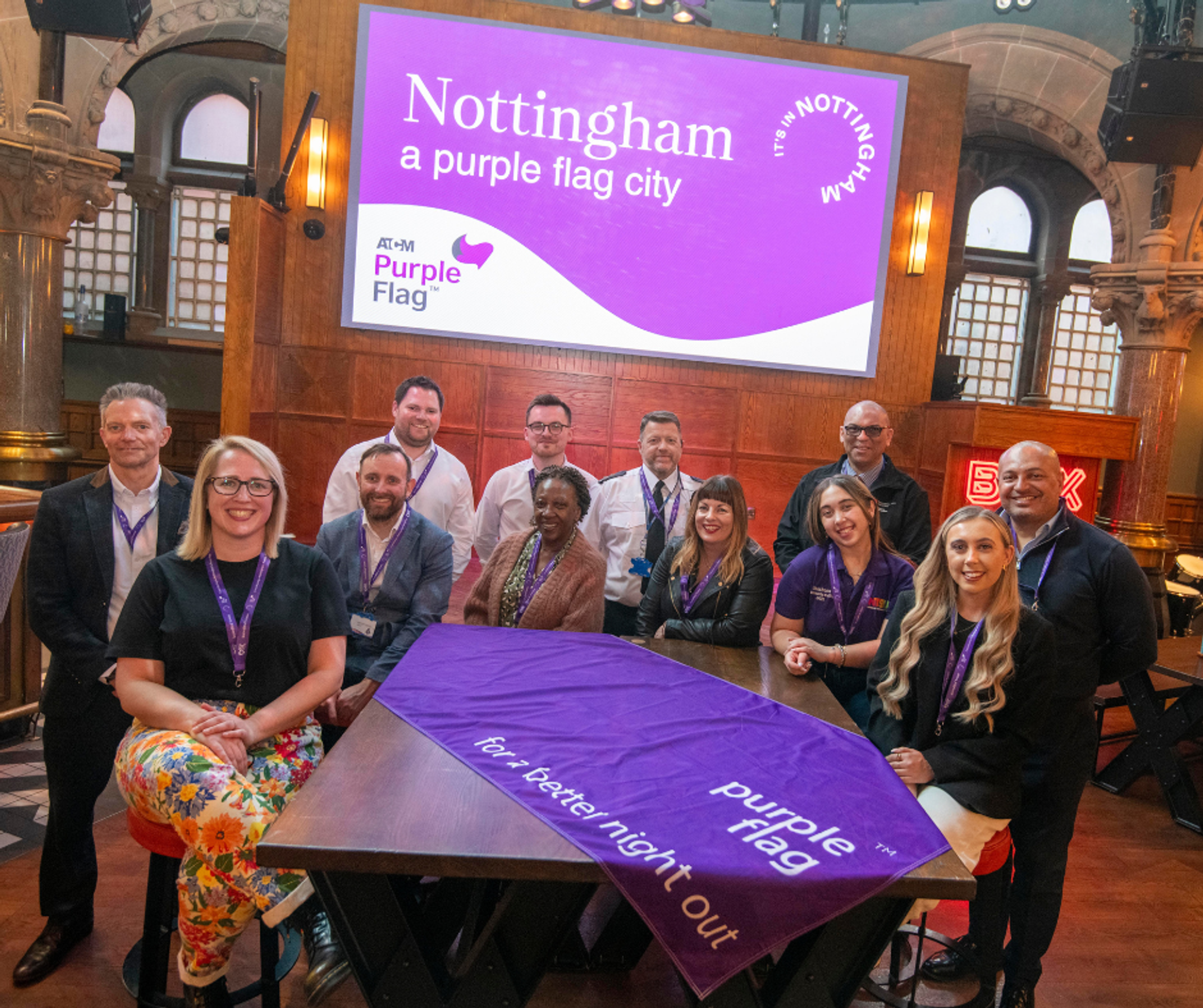 A group of people around a table smiling at the camera. There is a purple flag draped across the table reading 'purple flag: for a better night out', and a screen that reads 'Nottingham: a purple flag city'