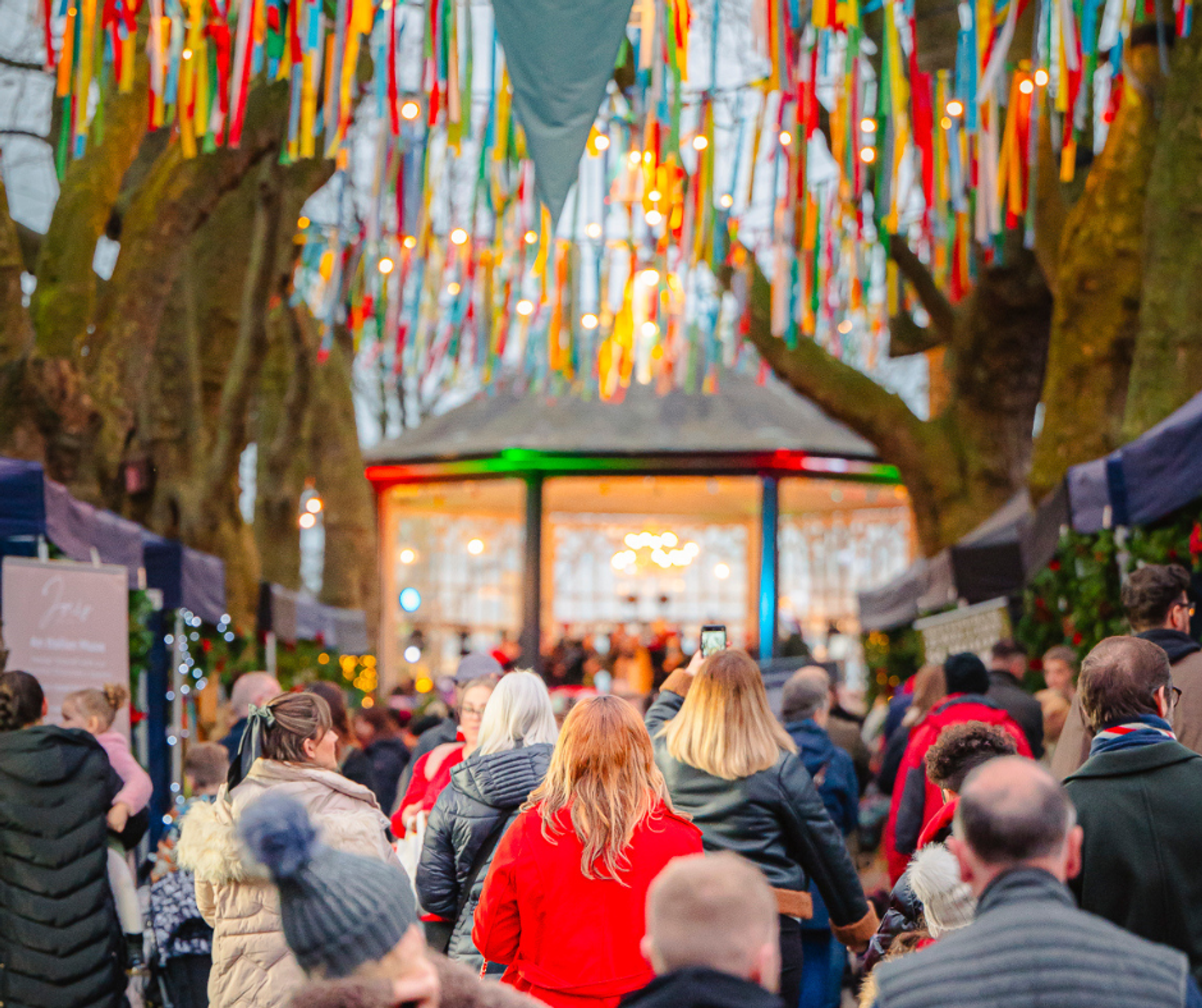 Visitors walk towards the bandstand at Nottingham Castle during the Christmas Market, with colourful ribbons and festoon lights above them.