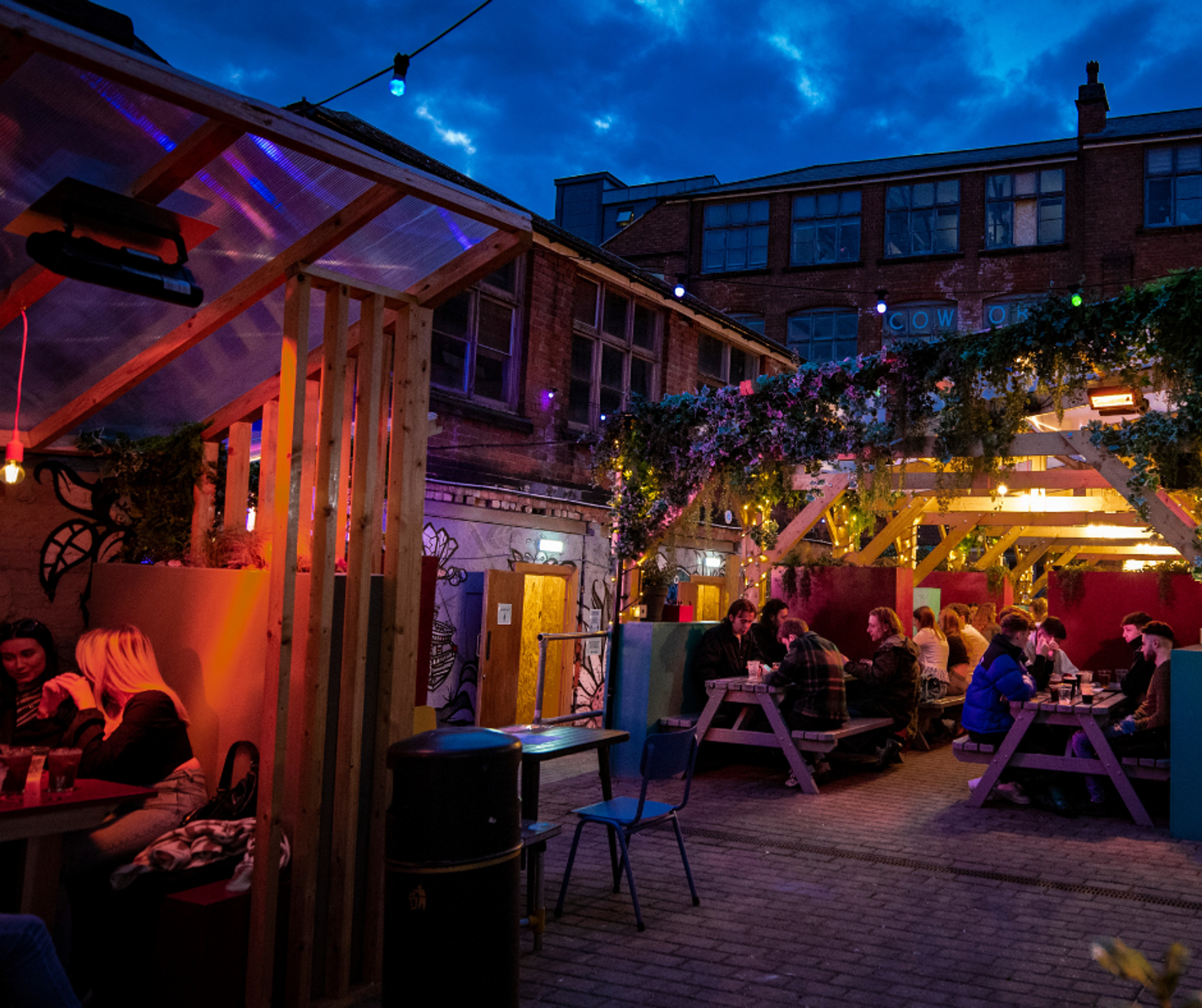 People in the outdoor courtyard area of a pub at night with seating, lighting and plants.